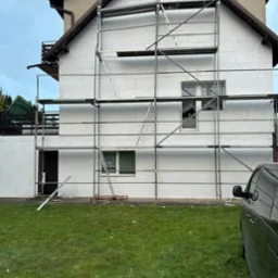 Dom w trakcie ocieplania, ściany pokryte styropianem, rusztowanie metalowe, widoczny fragment samochodu. Prace elewacyjne w toku.