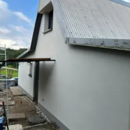 Świeżo otynkowana, jasnoszara elewacja domu z metalowym dachem, widok z boku. Widoczne rusztowanie i fragment chodnika z kostki brukowej.