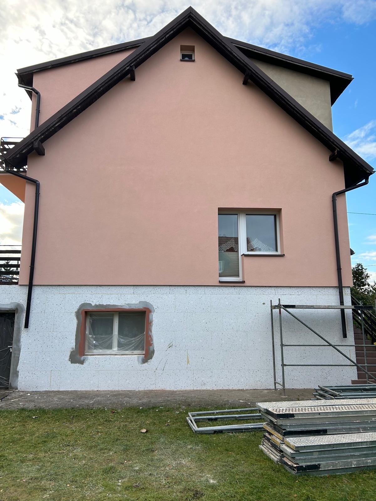 Różowa elewacja domu jednorodzinnego z widocznym ociepleniem styropianem na dole i rusztowaniem. Okno zabezpieczone folią. Częściowo ocieplony budynek.