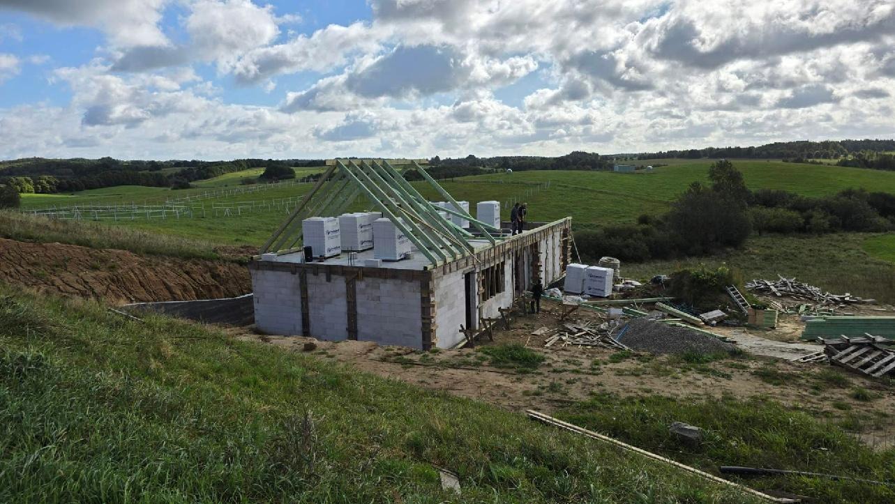 Budowa domu jednorodzinnego z bloczków, z widoczną konstrukcją dachu i materiałami budowlanymi na tle zielonego krajobrazu.