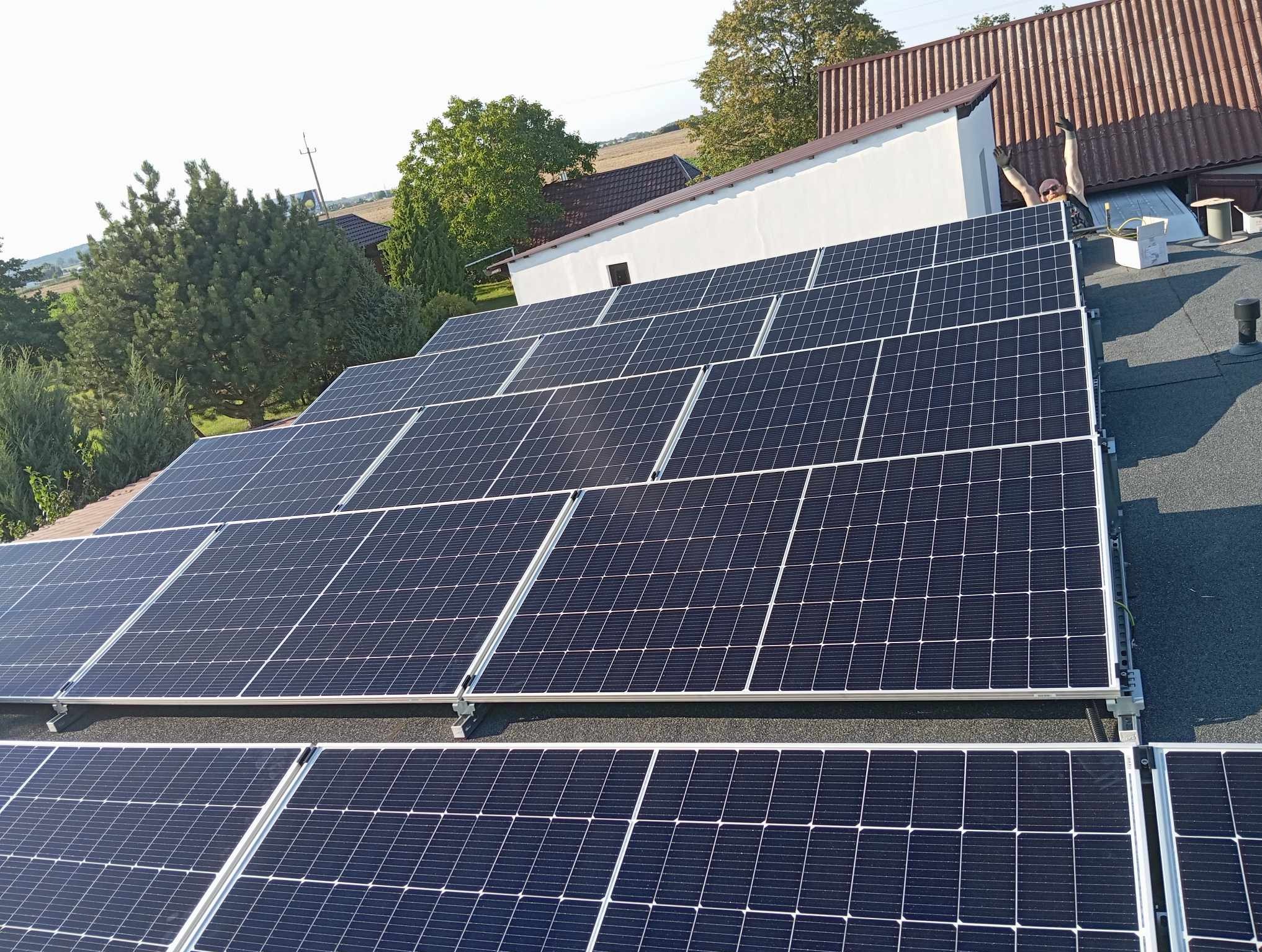 Instalacja paneli fotowoltaicznych na dachu budynku, widok z góry. Na drugim planie budynki gospodarcze i drzewa. Instalator z podniesionymi rękoma.