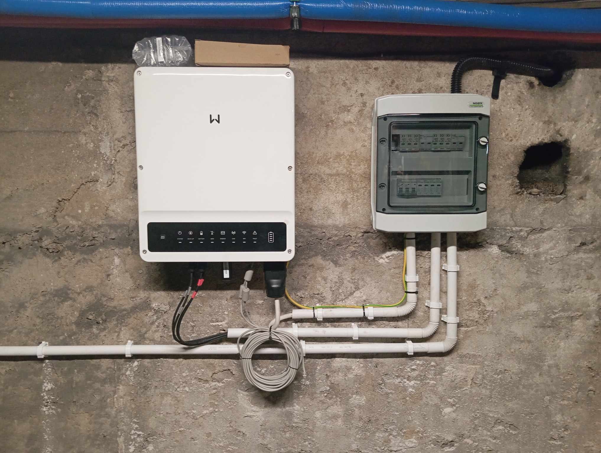 Biały inwerter fotowoltaiczny z symbolem 'M' i skrzynka elektryczna na surowej, betonowej ścianie. Widoczne kable i rury instalacyjne. Instalacja w piwnicy.