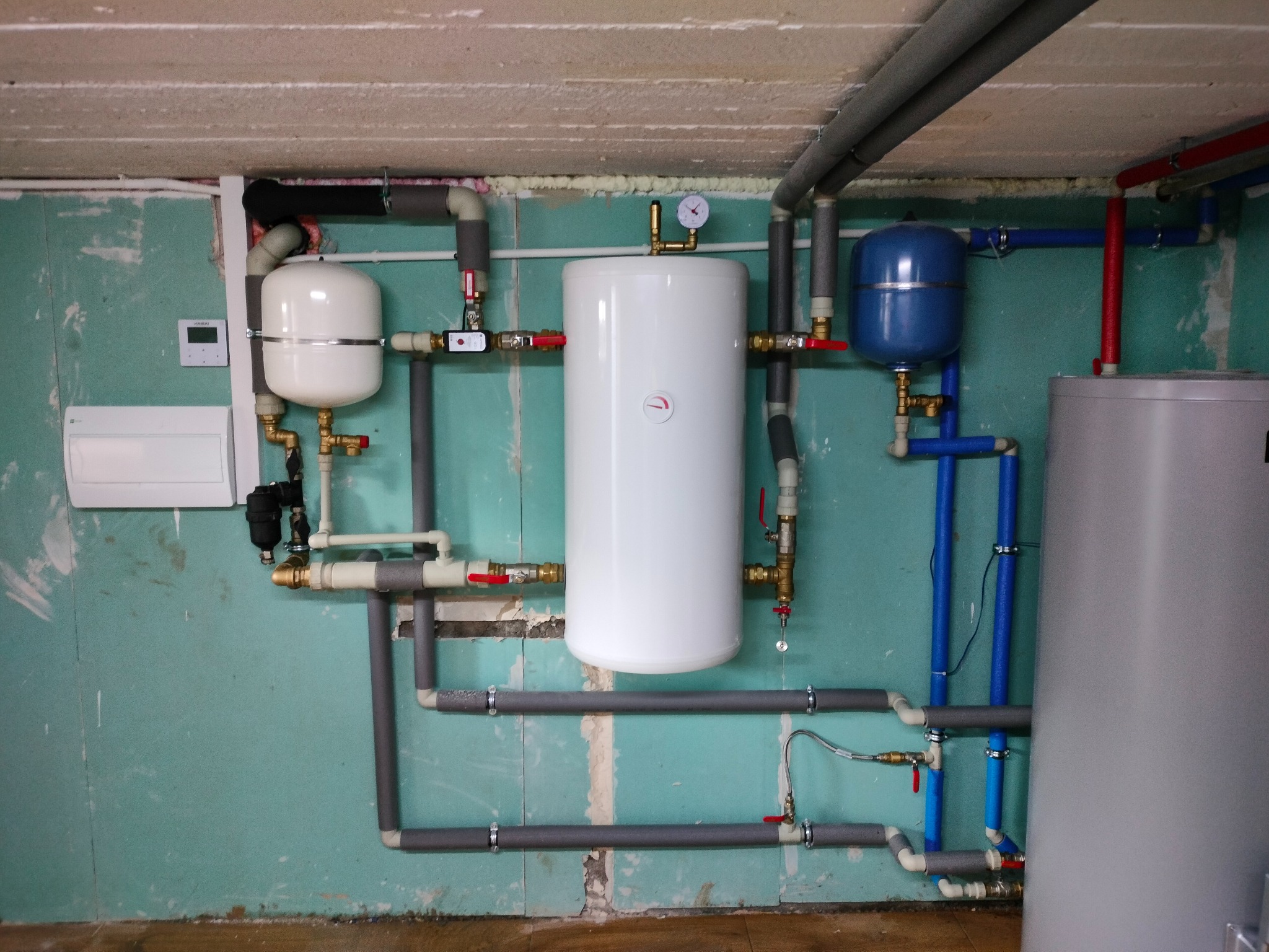 Kompletna instalacja grzewcza z białym zasobnikiem ciepłej wody, niebieskim naczyniem przeponowym i szarymi rurami w piwnicy z surowymi ścianami. Widoczny manometr i zawory.