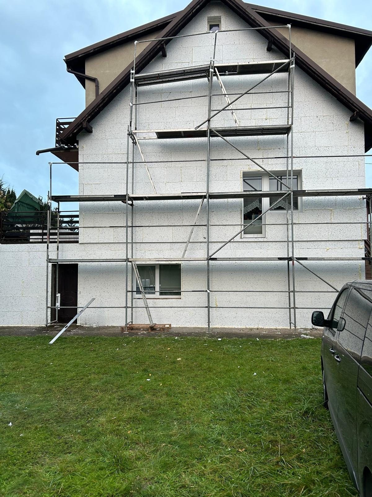 Dom w trakcie ocieplania, ściany pokryte styropianem, rusztowanie metalowe, widoczny fragment samochodu. Prace elewacyjne w toku.