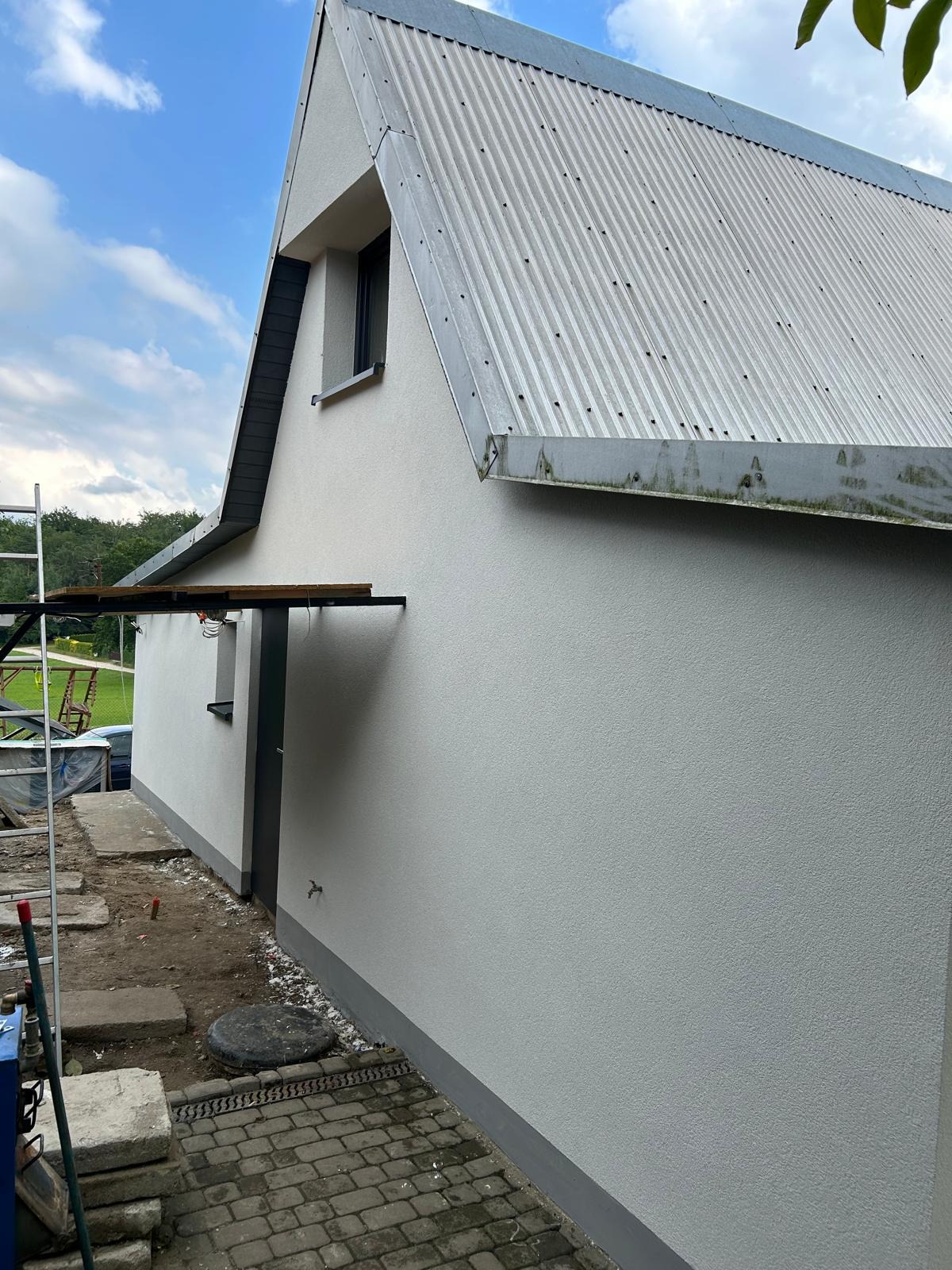 Świeżo otynkowana, jasnoszara elewacja domu z metalowym dachem, widok z boku. Widoczne rusztowanie i fragment chodnika z kostki brukowej.