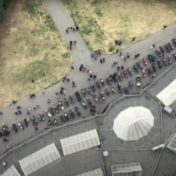 Widok z góry na zgromadzenie motocykli i ludzi na parkingu i ścieżce, obok drzew i budynku z przeszklonym dachem.