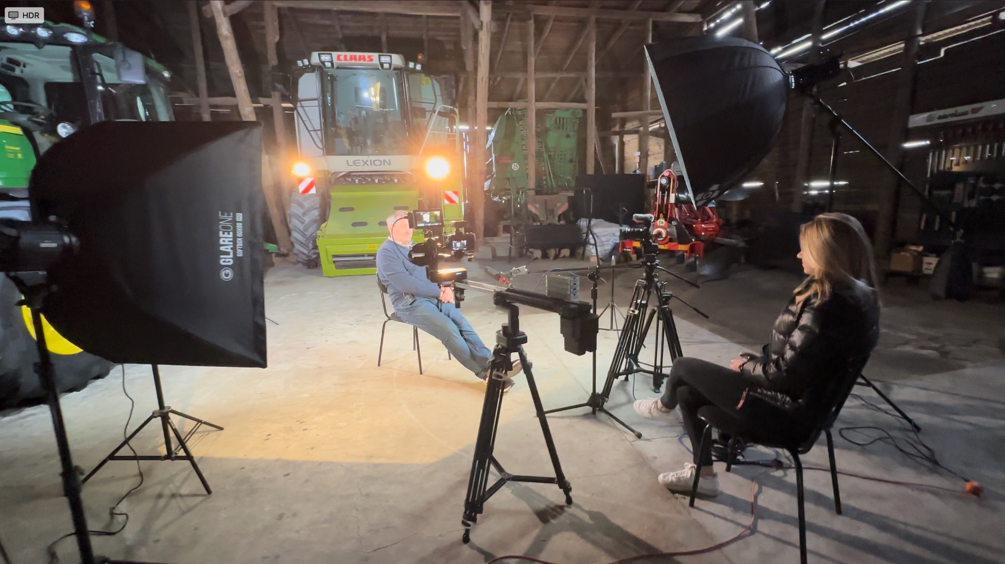 Plan filmowy w stodole: mężczyzna siedzi na krześle przed kombajnem, oświetlony softboxami, kobieta obserwuje z boku, kamery na statywach, suwnica kamerowa.