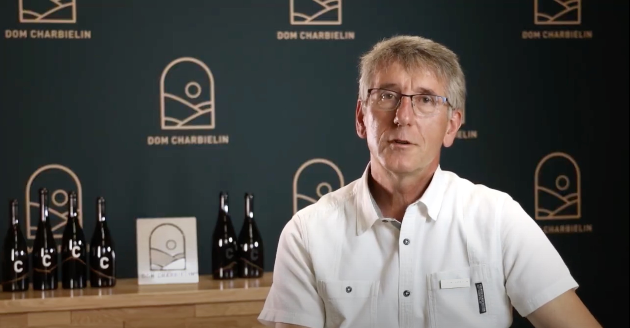 Mężczyzna w białej koszuli prezentuje się na tle ściany z logo Dom Charbielin i butelkami wina z logo firmy.