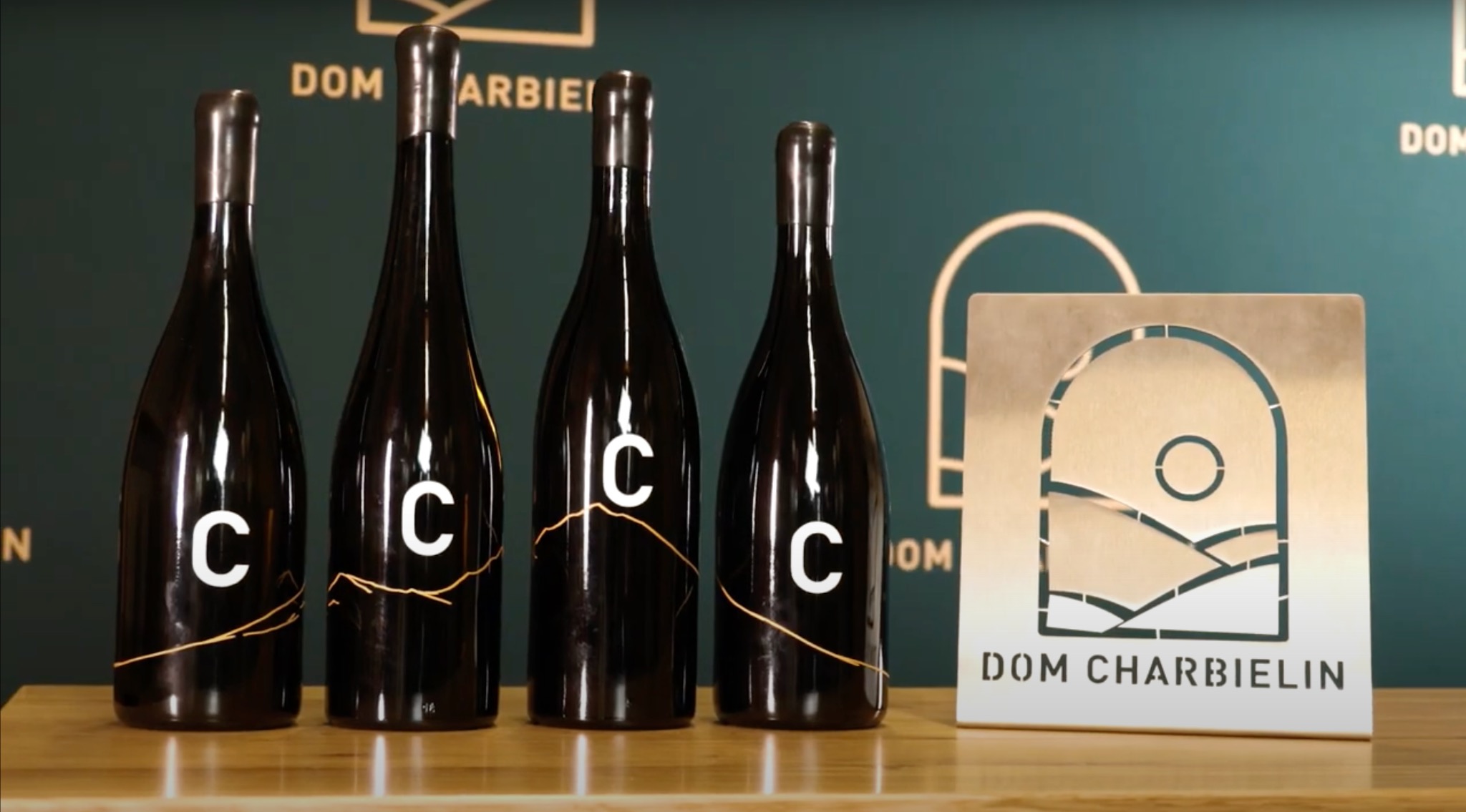 Cztery butelki ciemnego wina z białym logo 'C' i złotymi pęknięciami, obok metalowa tabliczka z wyciętym krajobrazem i napisem 'DOM CHARBIELIN' na drewnianym blacie i zielonym tle.