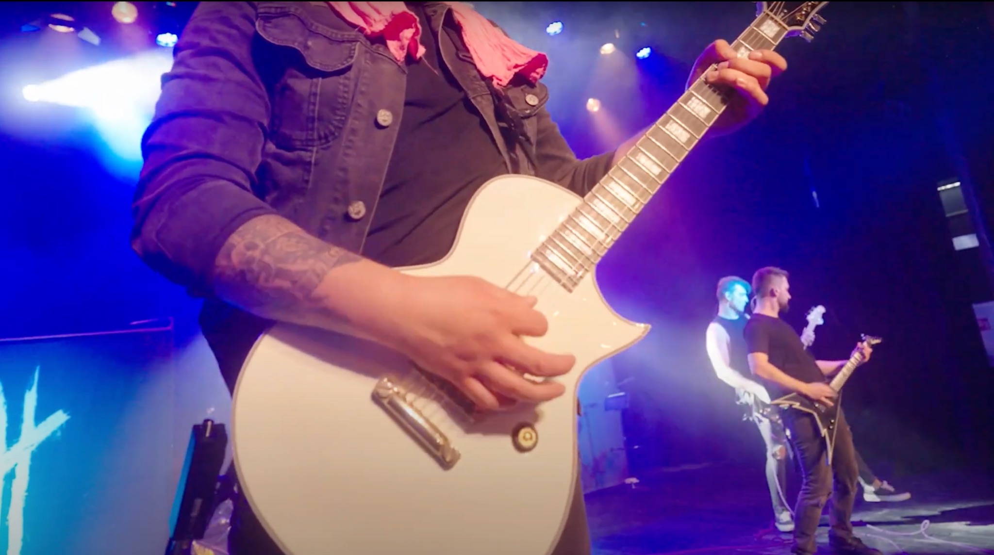 Gitarzysta z tatuażem na ramieniu gra na białej gitarze elektrycznej podczas koncertu, w tle widoczni inni muzycy i oświetlenie sceniczne.