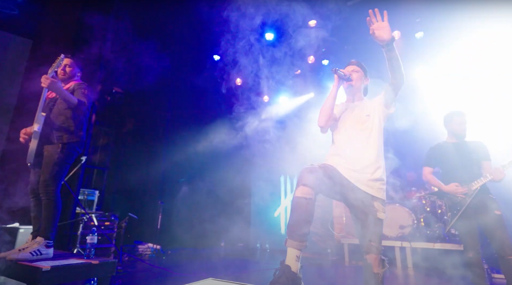 Zespół muzyczny na scenie w trakcie występu, wokalista z podniesioną ręką śpiewa do mikrofonu, w tle gitarzyści i perkusja, oświetlenie sceniczne i efekty dymne.