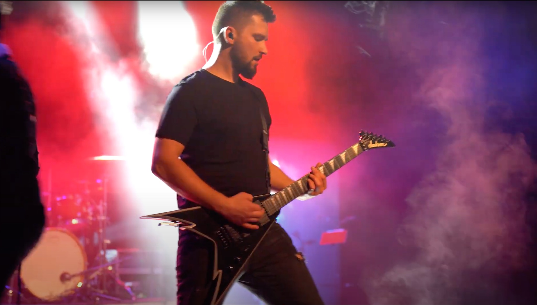 Gitarzysta gra na czarnej gitarze typu V na scenie, oświetlonej czerwonym i niebieskim światłem, z widoczną perkusją w tle.