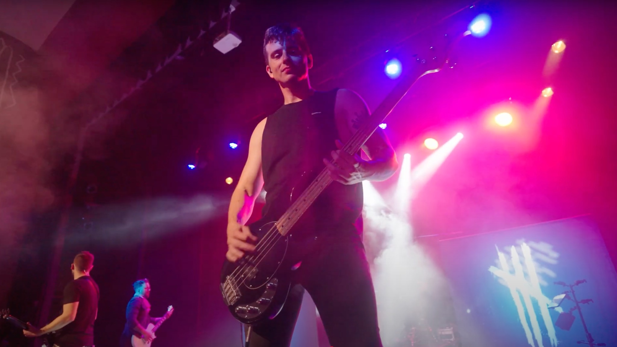 Basista gra na scenie w intensywnym oświetleniu, widoczni również inni muzycy w tle, scena wypełniona dymem.