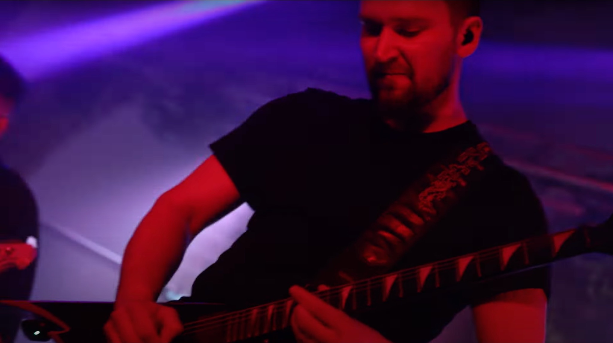Gitarzysta w czarnej koszulce gra na gitarze elektrycznej na scenie oświetlonej czerwonym i fioletowym światłem, z widocznym dymem i innym muzykiem w tle.
