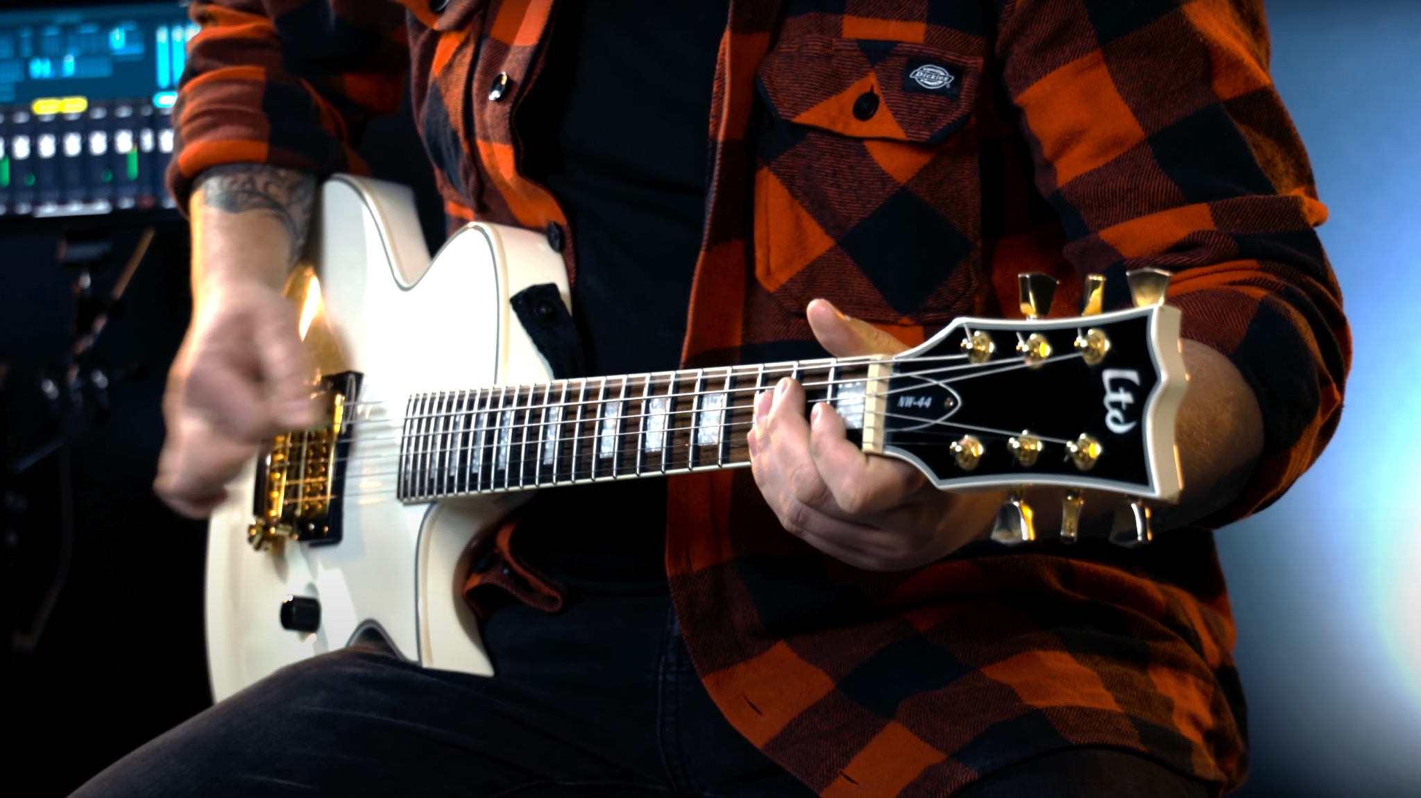 Mężczyzna w pomarańczowo-czarnej koszuli w kratę gra na białej gitarze elektrycznej w studio nagrań, widoczny mikser dźwięku w tle.