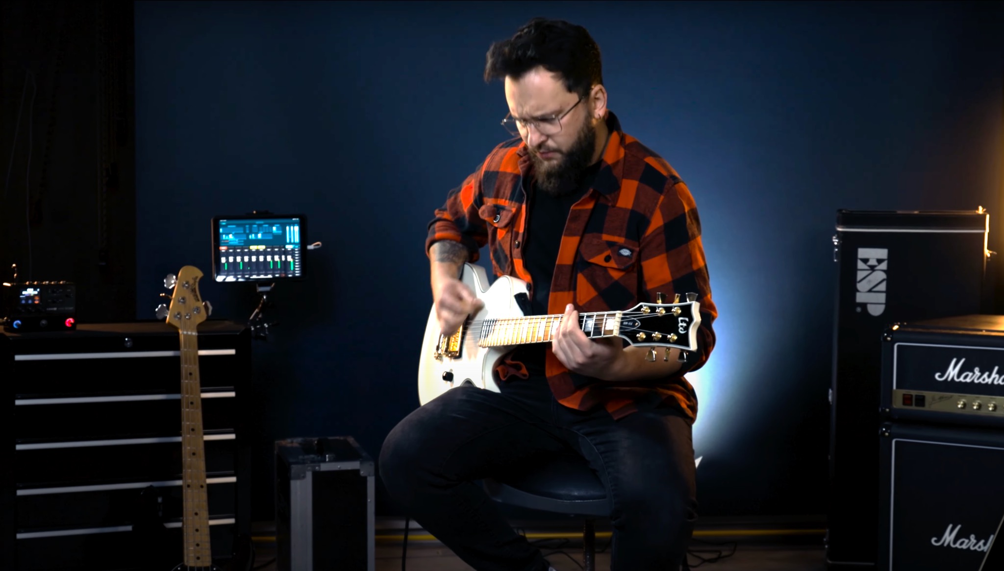 Brodaty gitarzysta w pomarańczowo-czarnej flanelowej koszuli gra na białej gitarze elektrycznej ESP, w tle wzmacniacze Marshall i ESP oraz tablet z oprogramowaniem do miksowania dźwięku.