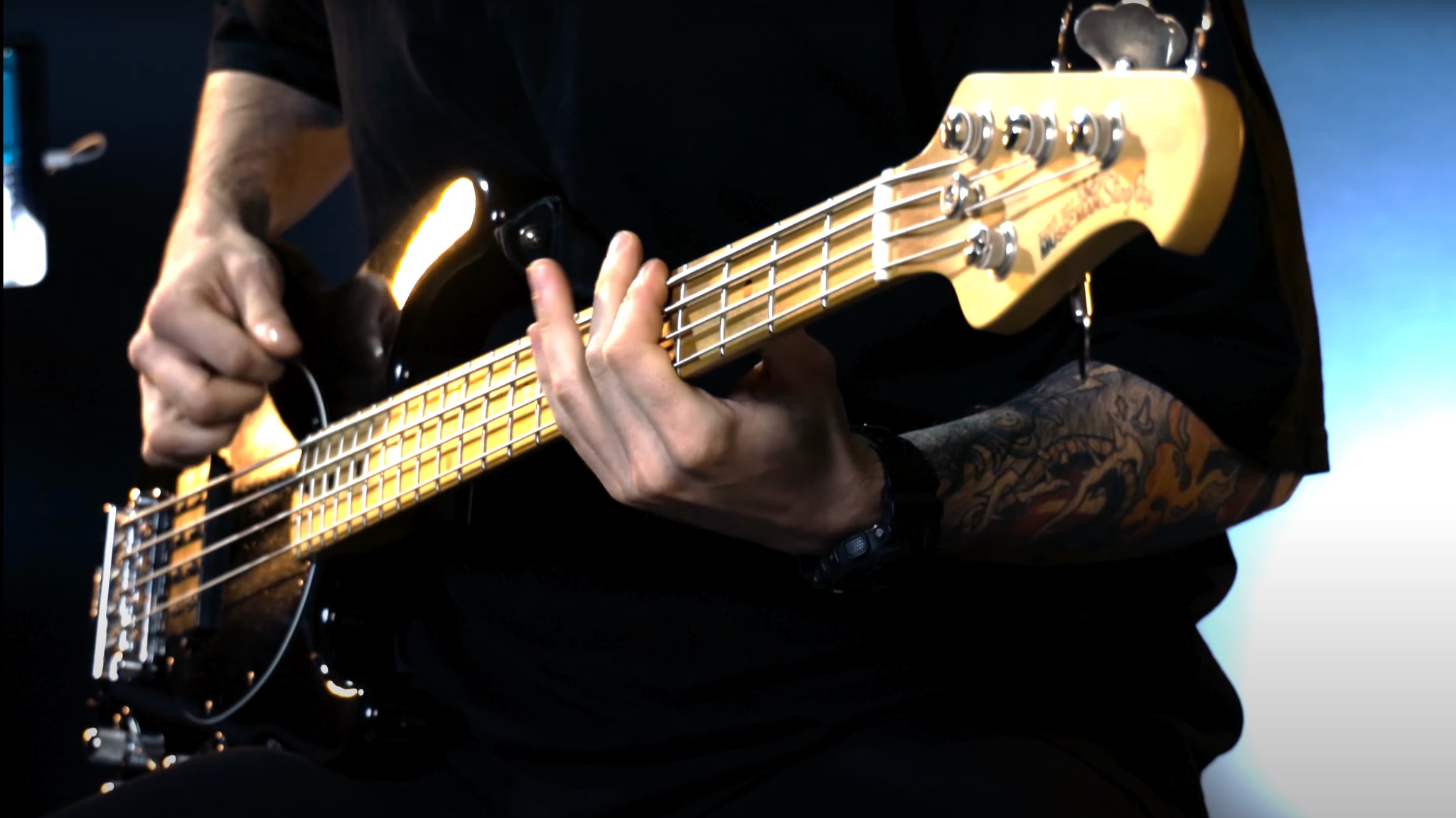 Mężczyzna z tatuażem na ramieniu gra na czarnej gitarze basowej z jasnym gryfem w studiu nagrań.