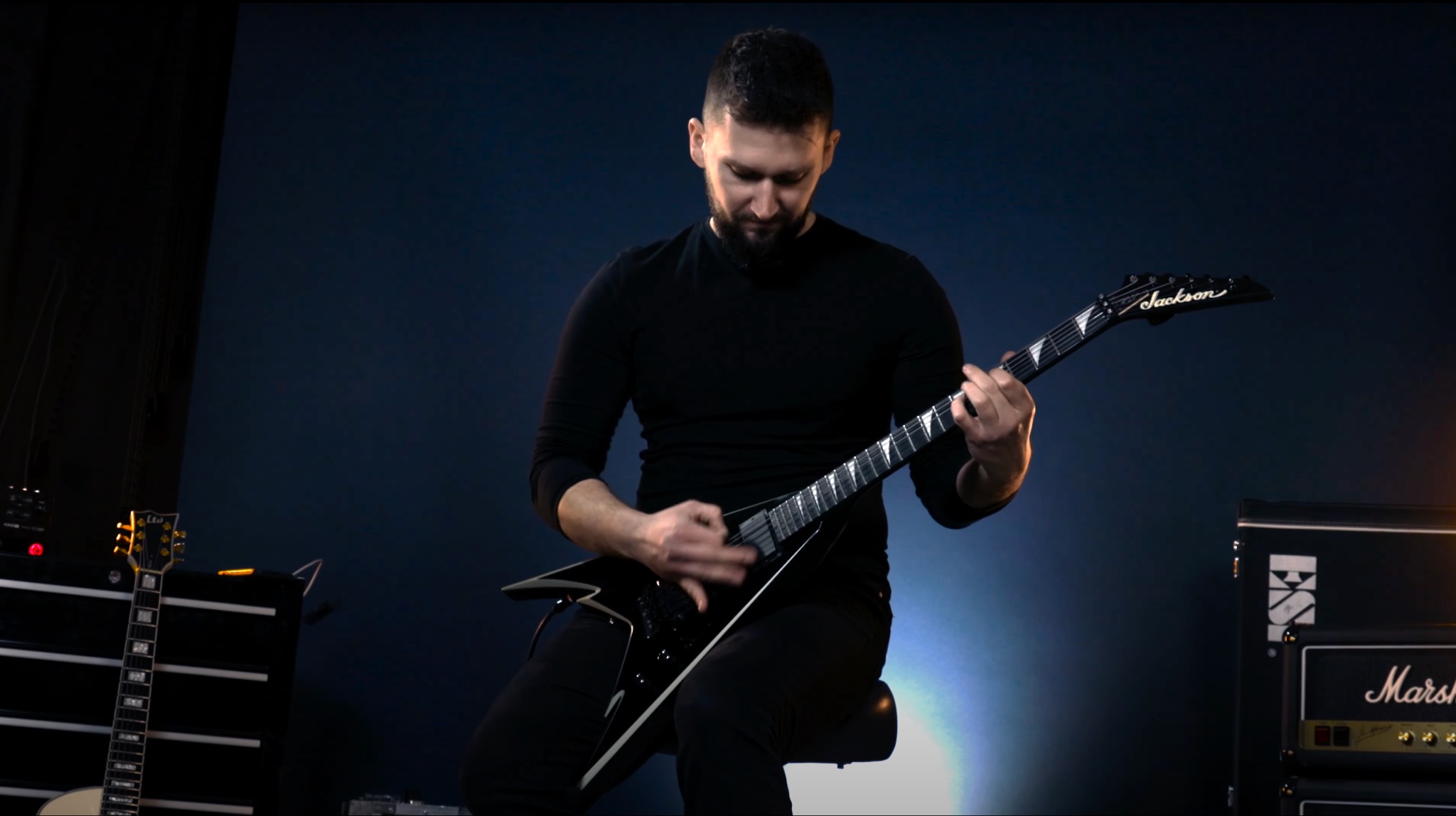Mężczyzna gra na czarnej gitarze elektrycznej Jackson V na tle wzmacniaczy i oświetlenia scenicznego.