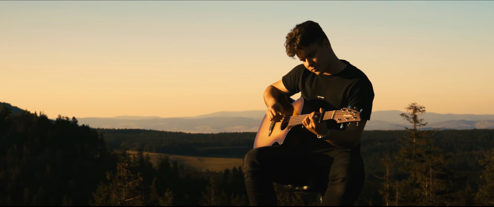 Młody mężczyzna gra na gitarze akustycznej na tle gór o zachodzie słońca, widoczne zalesione wzgórza i oddalona panorama.
