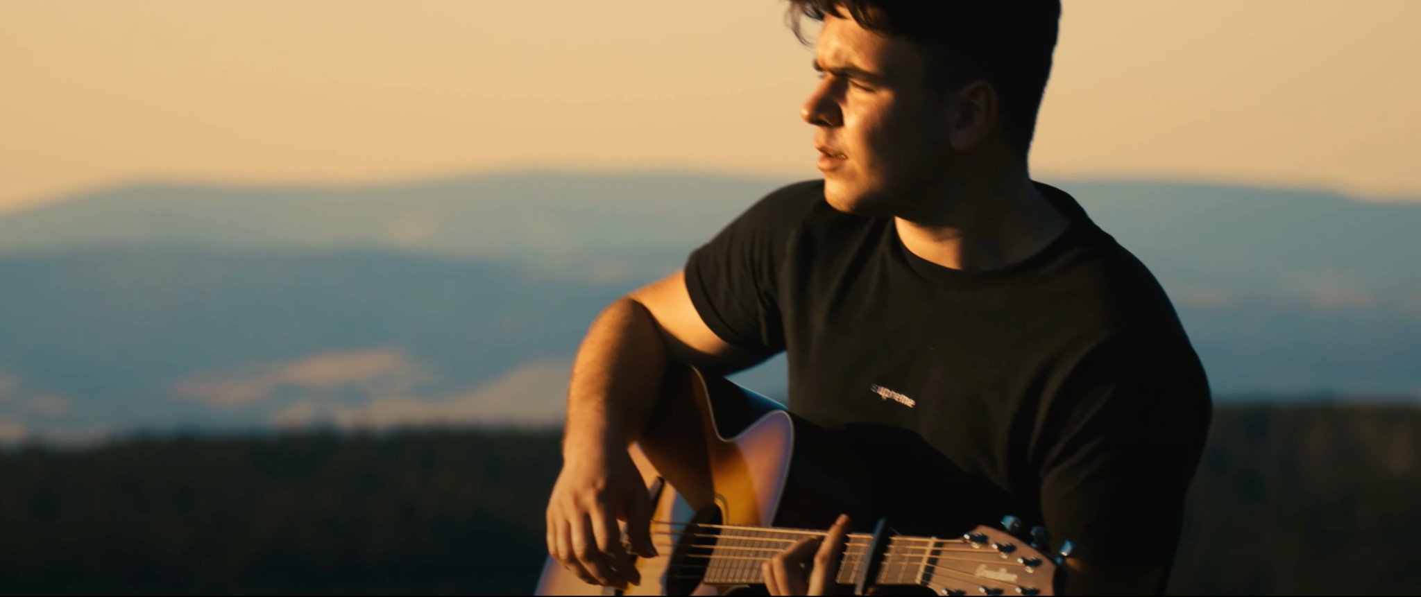 Młody mężczyzna gra na gitarze akustycznej na tle rozmytego krajobrazu górskiego o zachodzie słońca, uchwycony w ciepłym, naturalnym świetle.