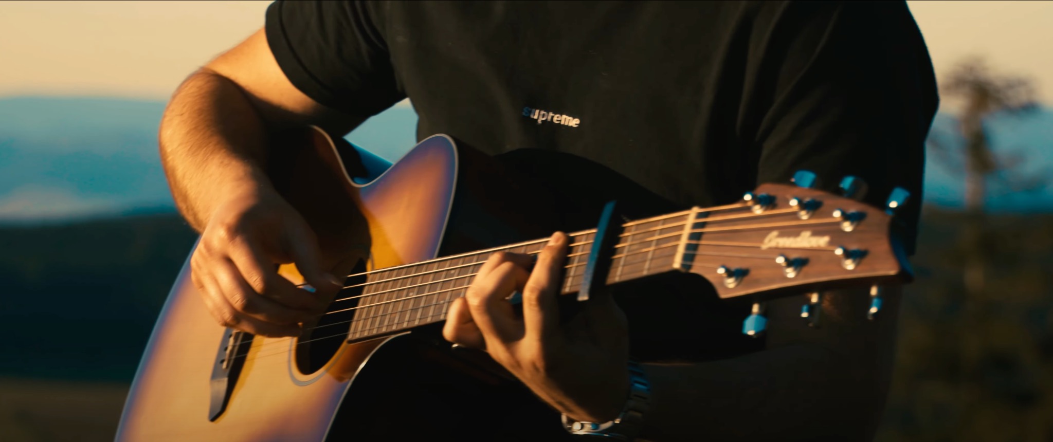 Mężczyzna grający na akustycznej gitarze z założonym kapodasterem na tle rozmytego krajobrazu górskiego o zachodzie słońca.