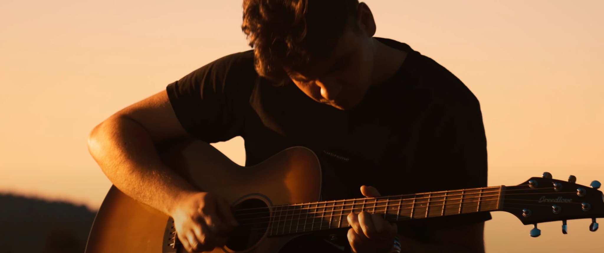 Młody mężczyzna gra na gitarze akustycznej Breedlove w plenerze, oświetlony ciepłym światłem zachodzącego słońca.