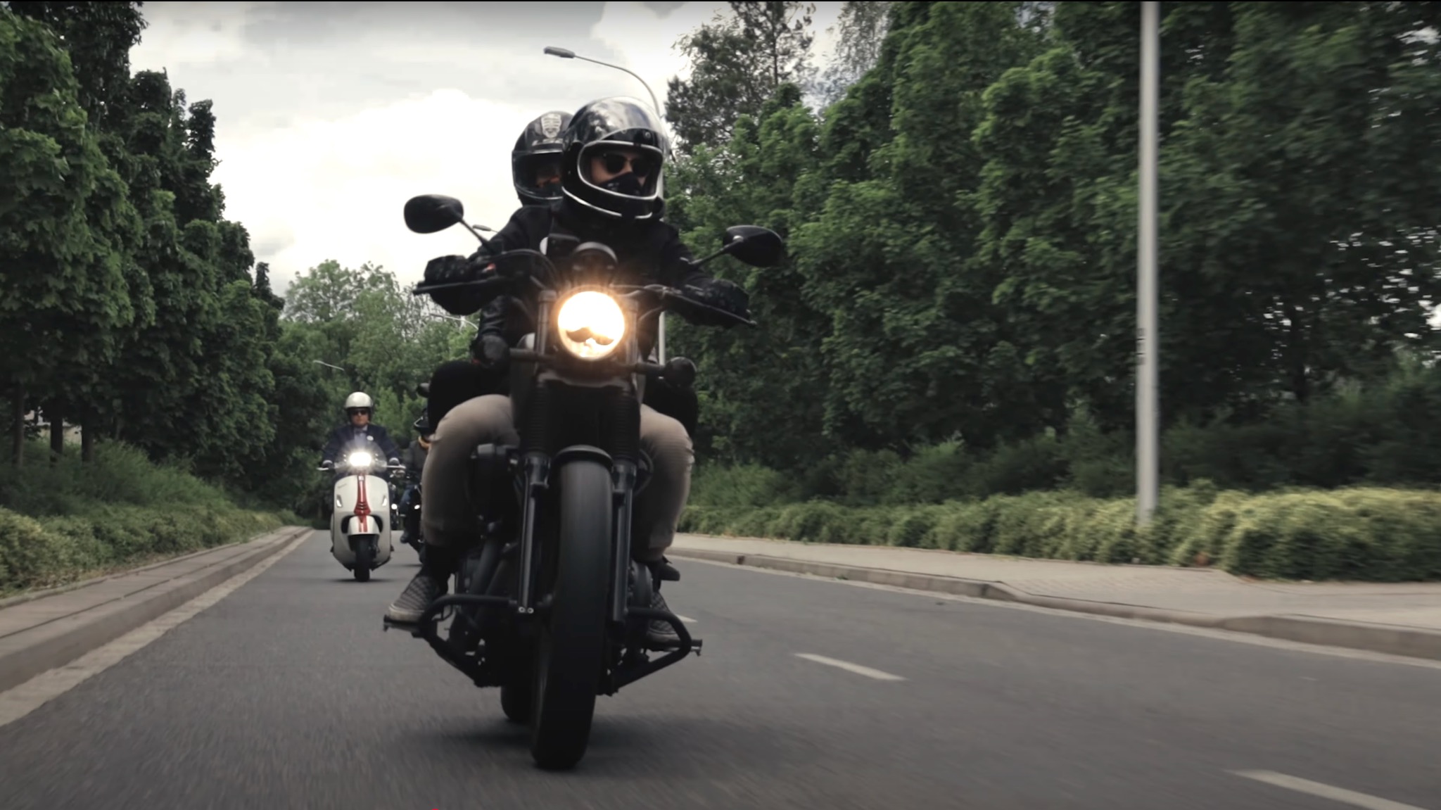 Kamerzysta filmuje z motocykla grupę osób jadących skuterami po asfaltowej drodze otoczonej zielenią.