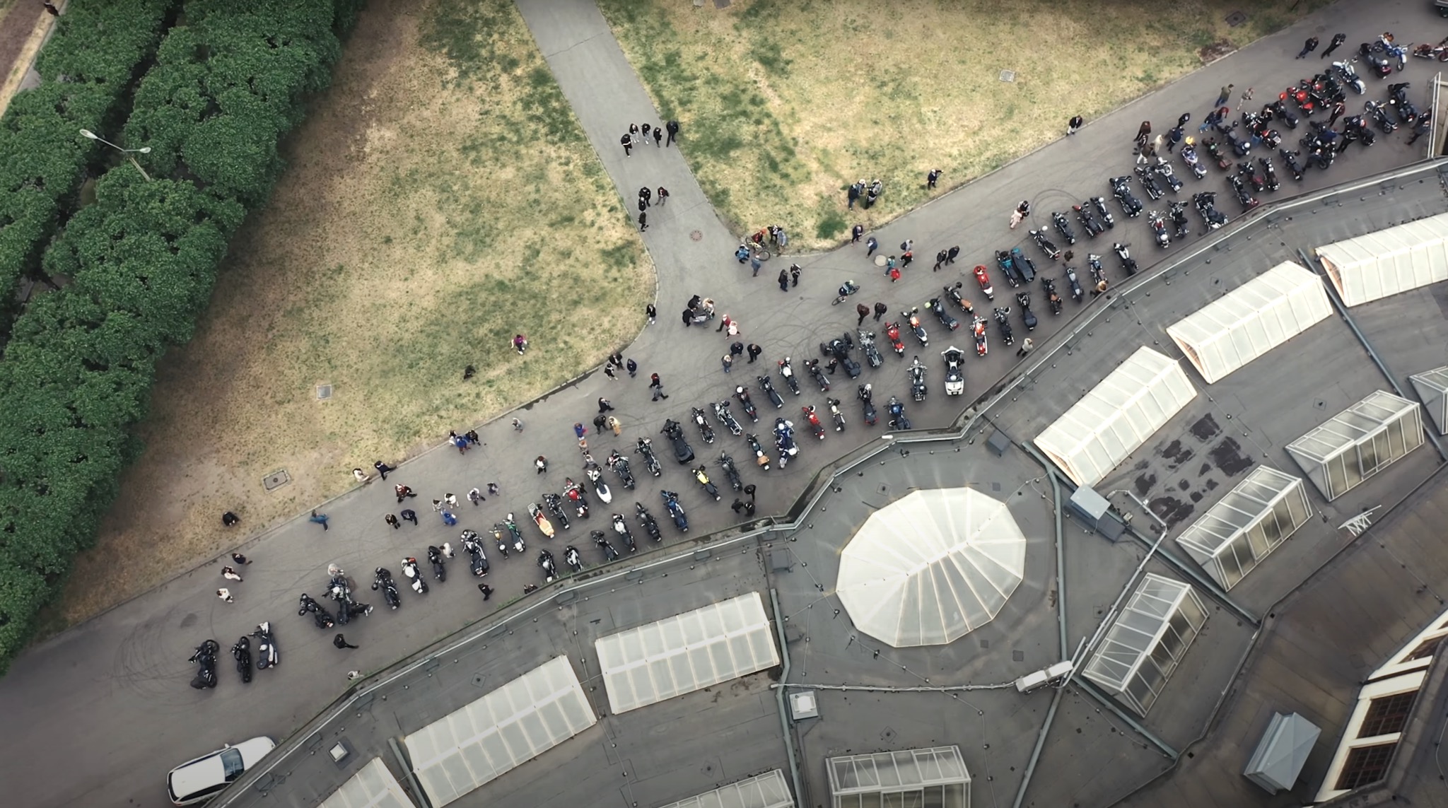 Widok z góry na zgromadzenie motocykli i ludzi na parkingu i ścieżce, obok drzew i budynku z przeszklonym dachem.