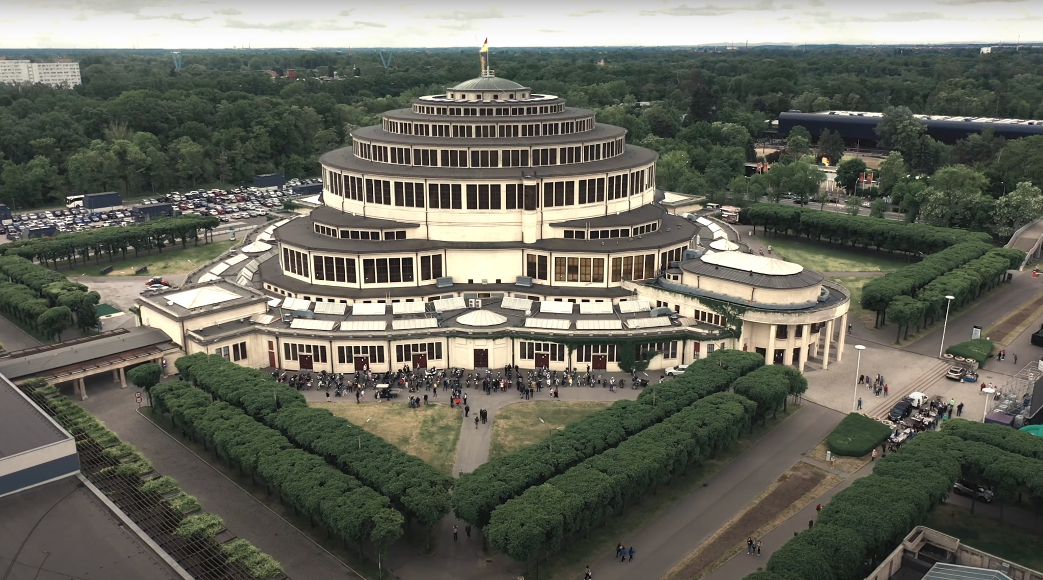 Panorama Hali Stulecia we Wrocławiu z lotu ptaka, otoczonej zielenią i parkingiem, widoczny tłum ludzi przed wejściem.