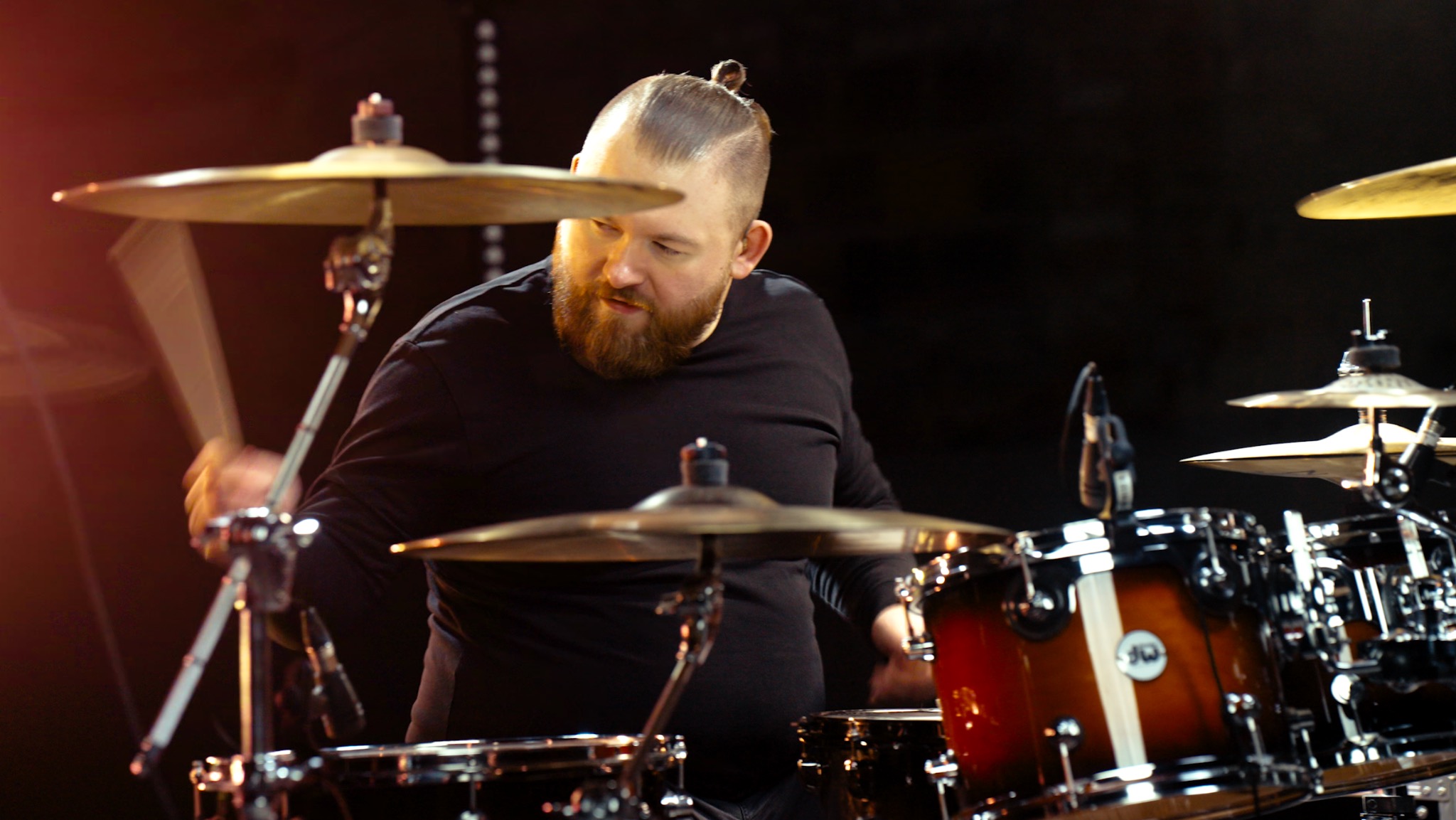 Perkusista z brodą i włosami spiętymi w kok gra na perkusji na scenie, ujęcie z bliska z widocznymi talerzami i bębnami.