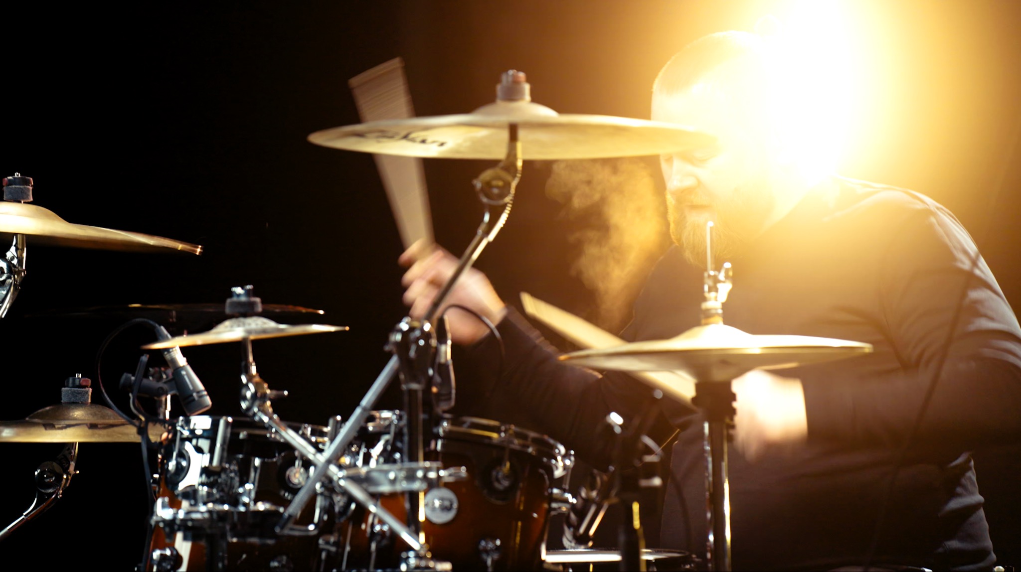 Perkusista z brodą gra na perkusji pod silnym światłem, uderzając pałeczkami w talerze, widoczny dym.