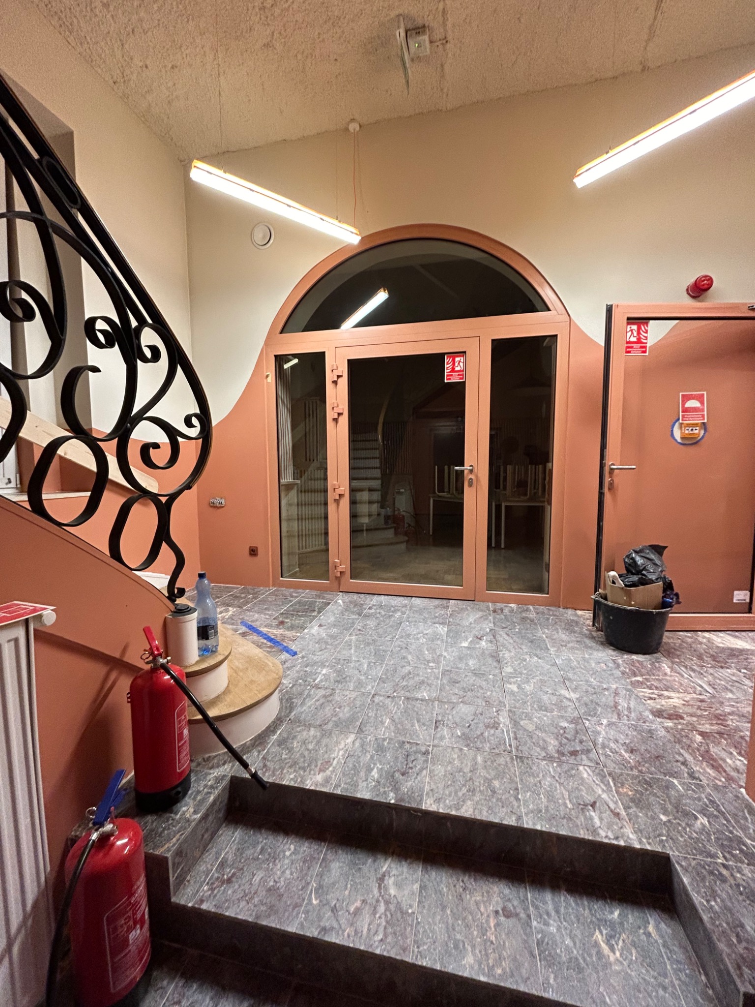 Korytarz w trakcie prac wykończeniowych: widoczne dwa gaśnice, schody z marmurowymi płytkami, drzwi z szybami oraz ślady po remoncie.