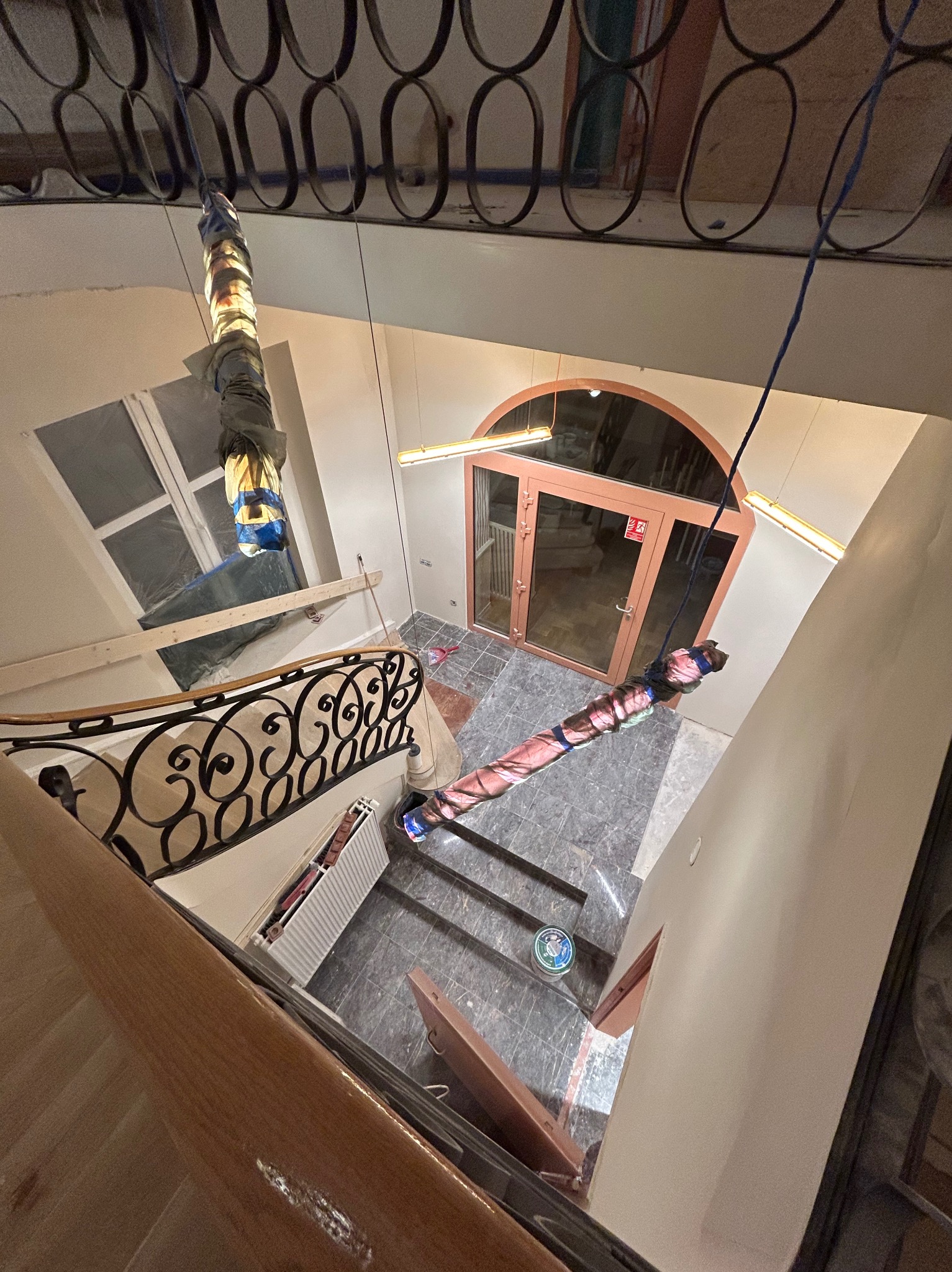 Klatka schodowa w trakcie prac zabezpieczających elementy przed zabrudzeniem, widoczne zakryte lampy i poręcze, okno zabezpieczone folią, oraz drzwi wejściowe z ościeżnicą.