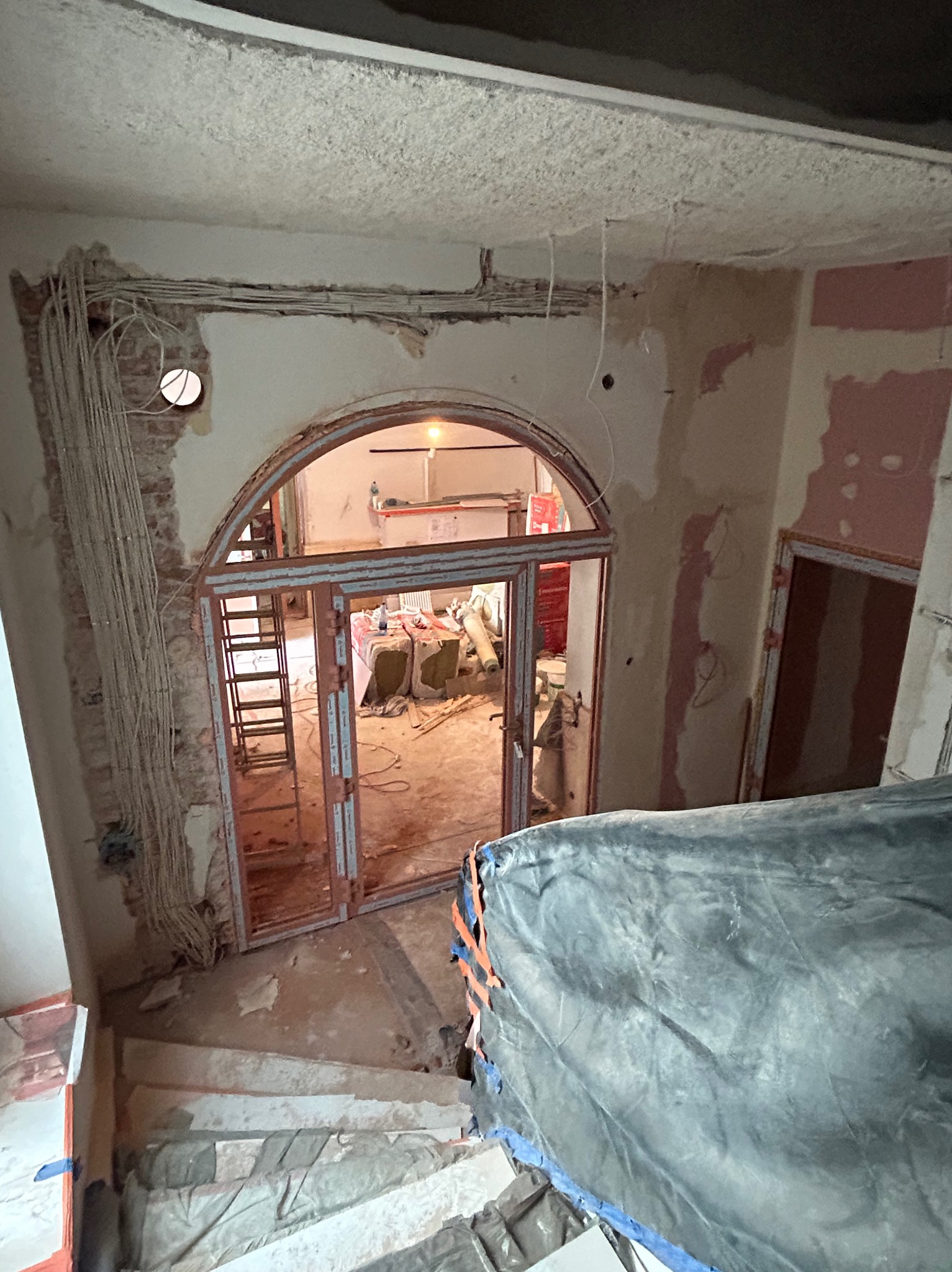 Wnętrze w trakcie prac wykończeniowych: odsłonięta cegła, instalacje elektryczne na ścianie, schody zabezpieczone folią, widok na pomieszczenie z drabiną i materiałami budowlanymi.