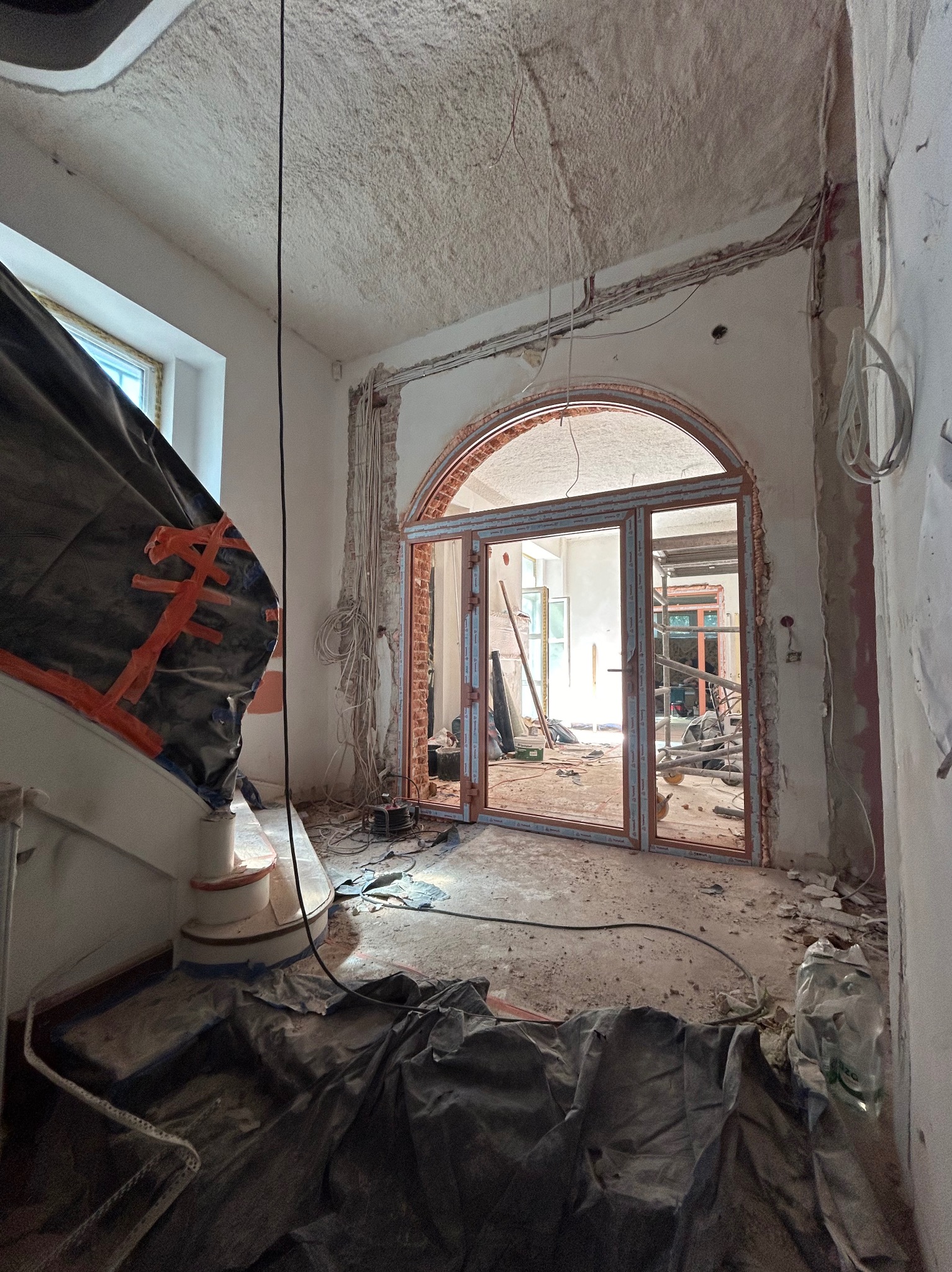 Korytarz w trakcie prac remontowych z widoczną konstrukcją łukowego otworu drzwiowego, zabezpieczonymi schodami oraz odsłoniętą instalacją elektryczną na ścianach i suficie.