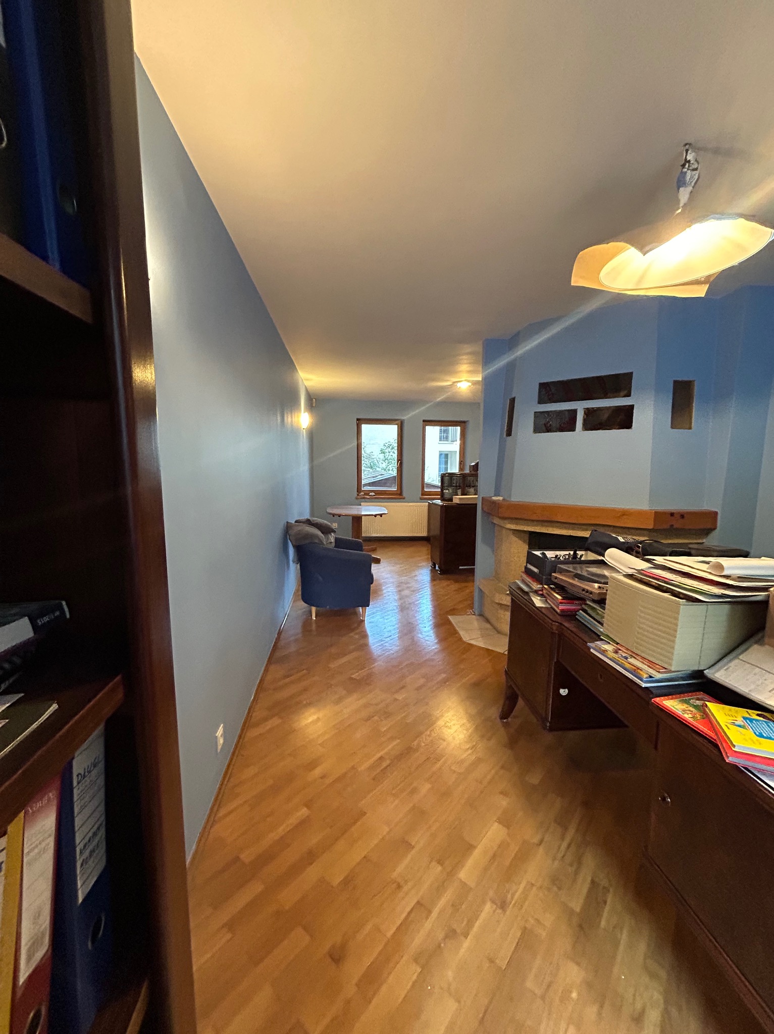 Wnętrze pokoju z niebieskimi ścianami, parkietem i kominkiem, widok z perspektywy regału z segregatorami.