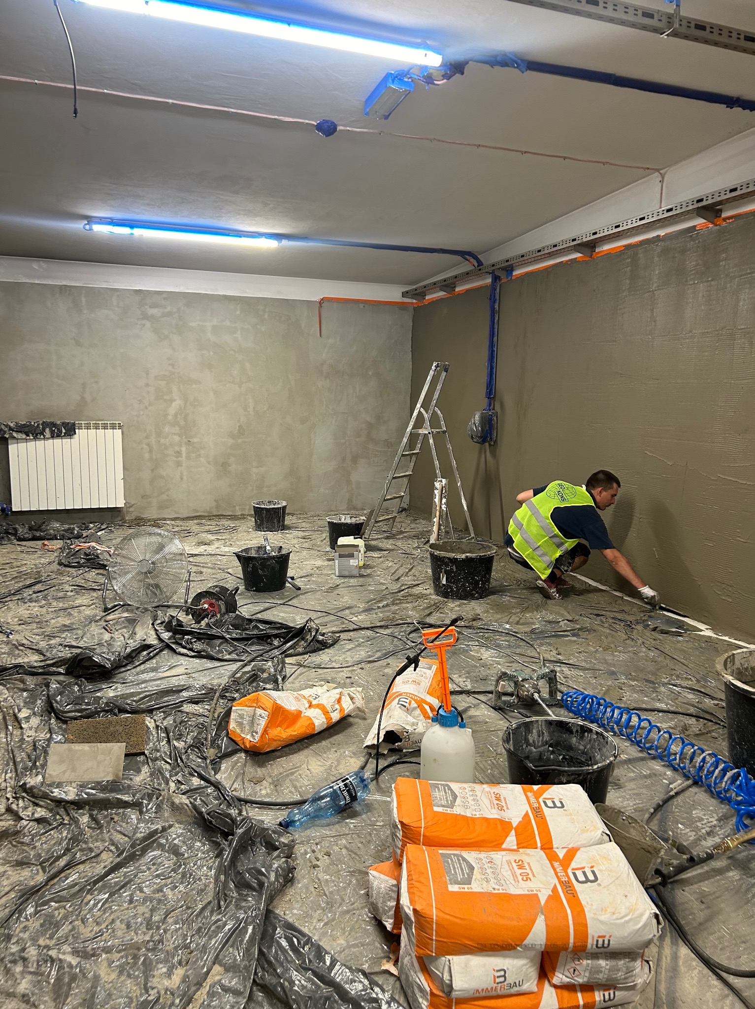 Pracownik w kamizelce odblaskowej aplikuje gładź na ścianę w pomieszczeniu przygotowanym do remontu, z rozłożoną folią ochronną, wiadrami, workami z gładzią i narzędziami.