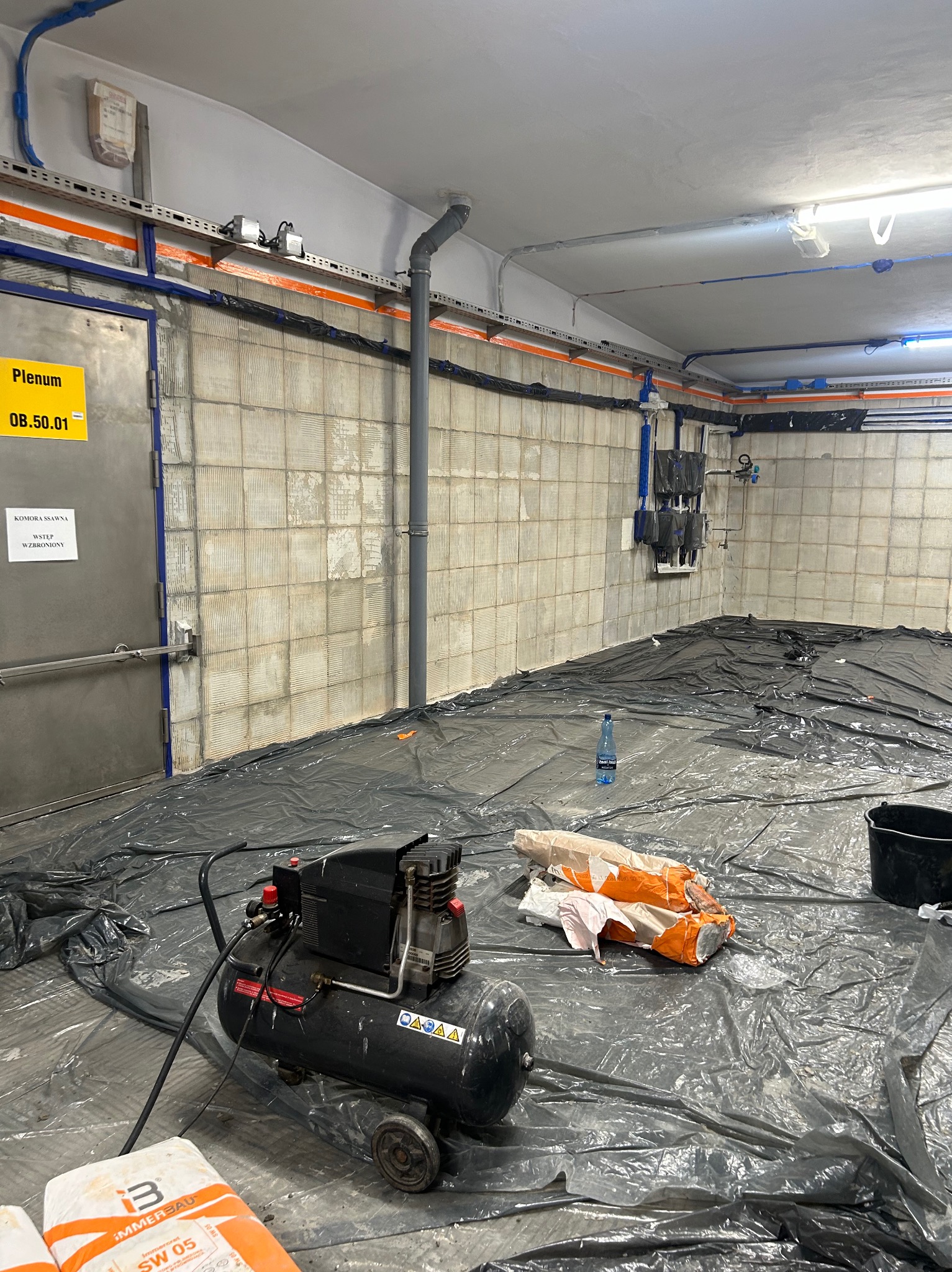 Przygotowania do prac w pomieszczeniu: ściany z płytkami wymagające obróbki, podłoga zabezpieczona folią, kompresor, worki z zaprawą i wiadro.