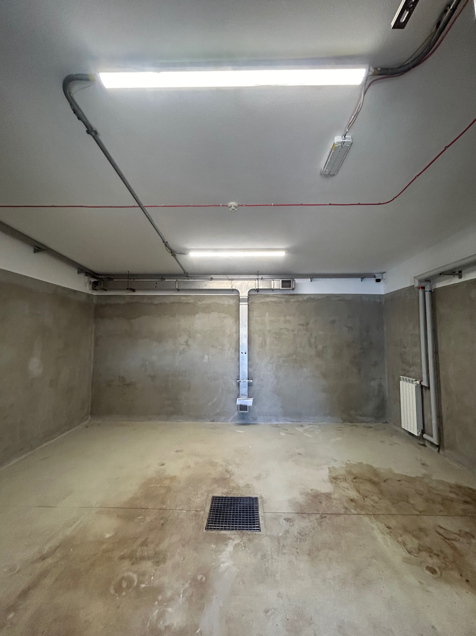Surowe wnętrze pomieszczenia z betonowymi ścianami, kratką ściekową w podłodze i instalacjami pod sufitem, gotowe do dalszych prac wykończeniowych.