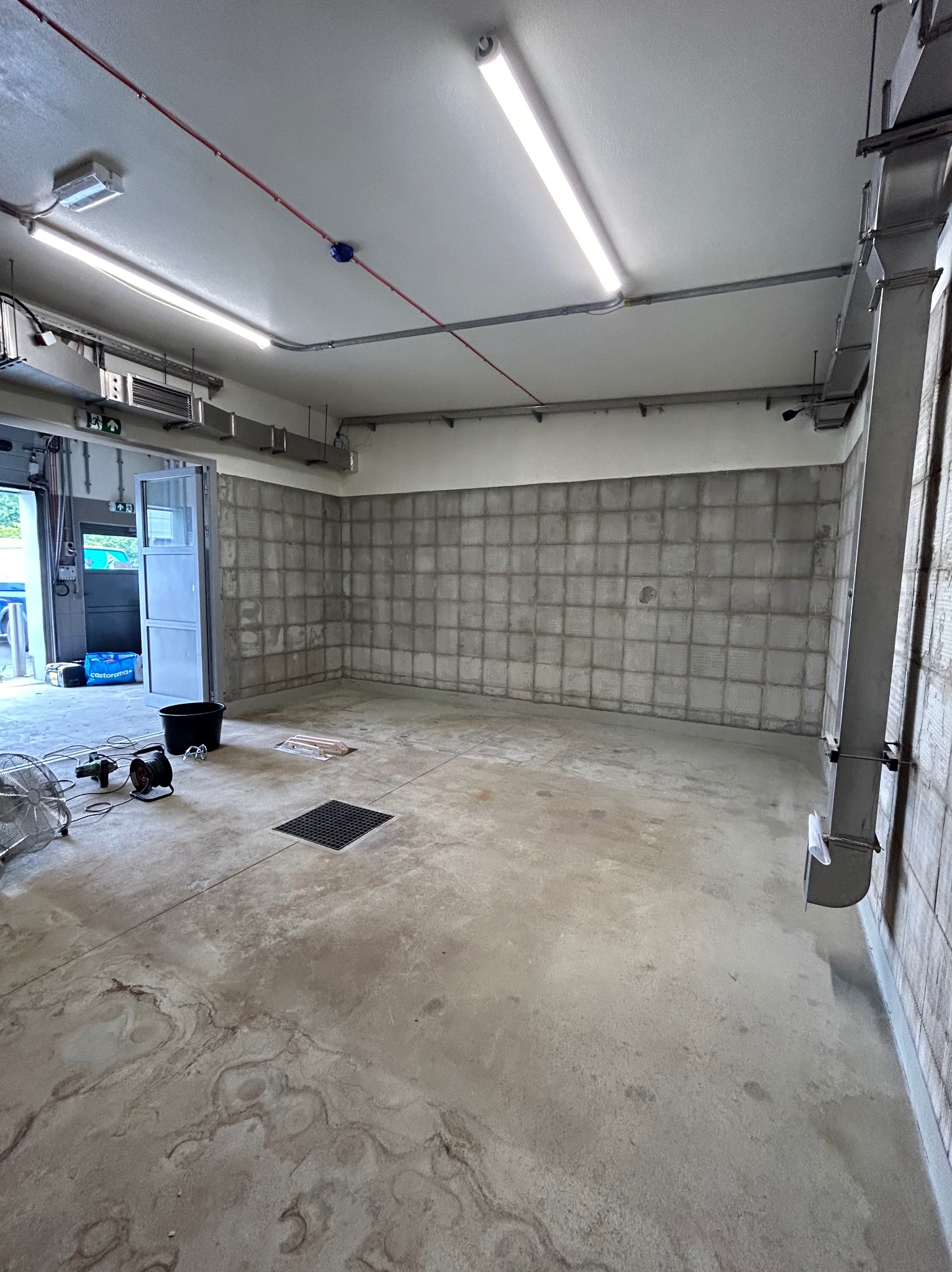 Przygotowane pomieszczenie biurowe przed pracami wykończeniowymi, z widoczną ścianą z surowego betonu w kwadraty, wentylacja pod sufitem, narzędzia i wiadro na podłodze.