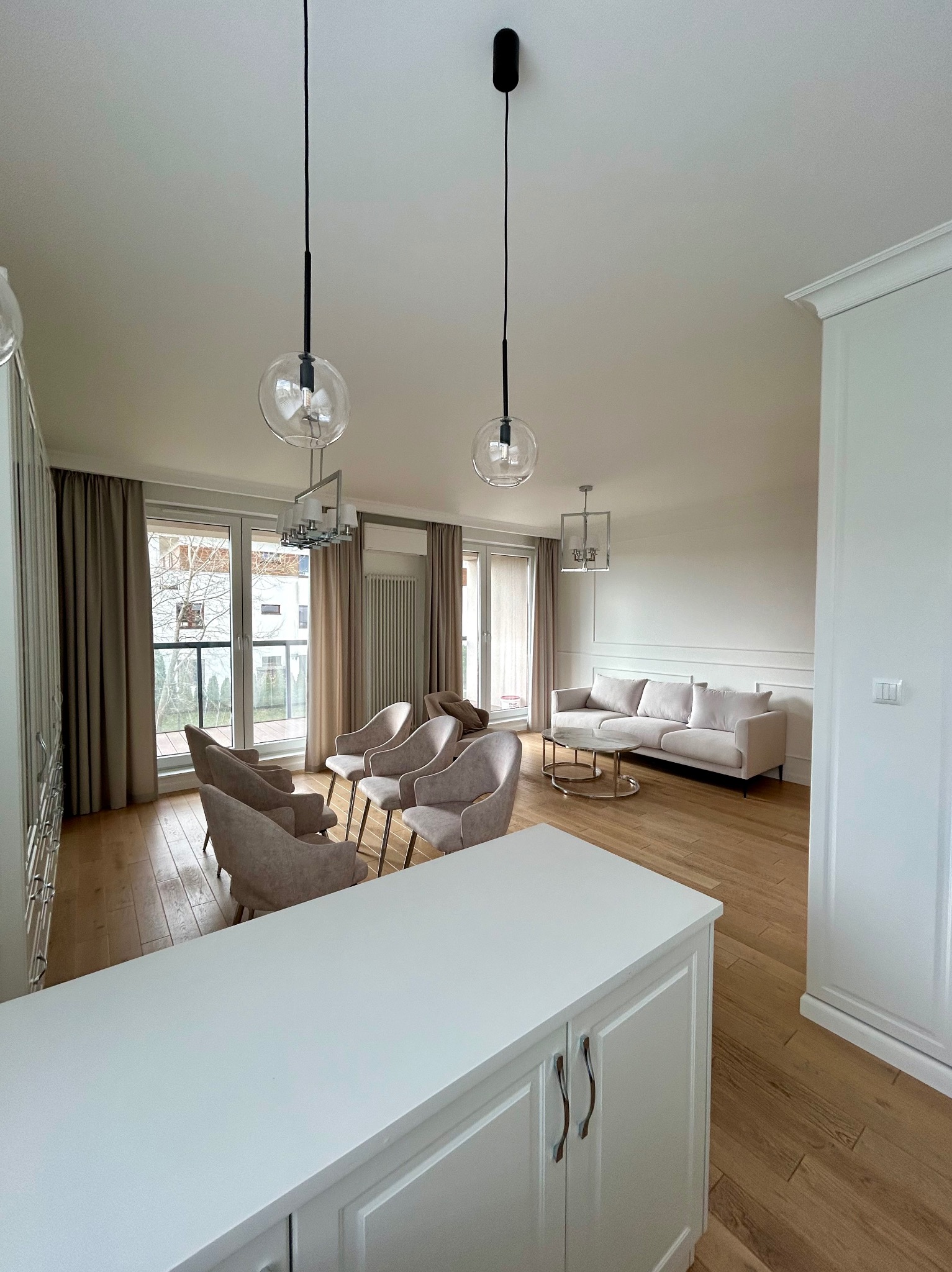 Jasne wnętrze salonu z drewnianą podłogą, beżowymi zasłonami i meblami w odcieniach bieli i beżu, oświetlone nowoczesnymi lampami wiszącymi z przezroczystymi kloszami.