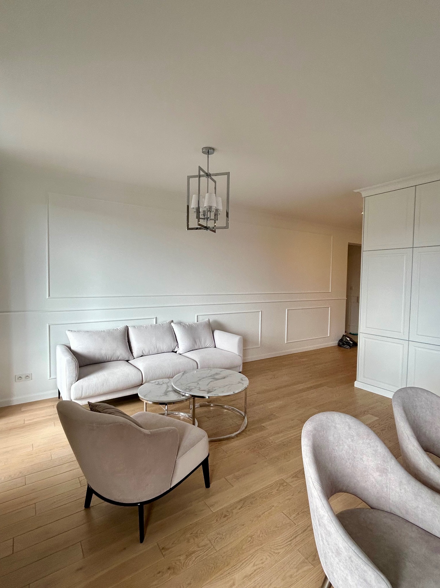 Jasne wnętrze salonu po remoncie, z drewnianą podłogą, białymi ścianami z dekoracyjnymi listwami, sofą, stolikami kawowymi z marmurowymi blatami, krzesłami i nowoczesną lampą wiszącą.