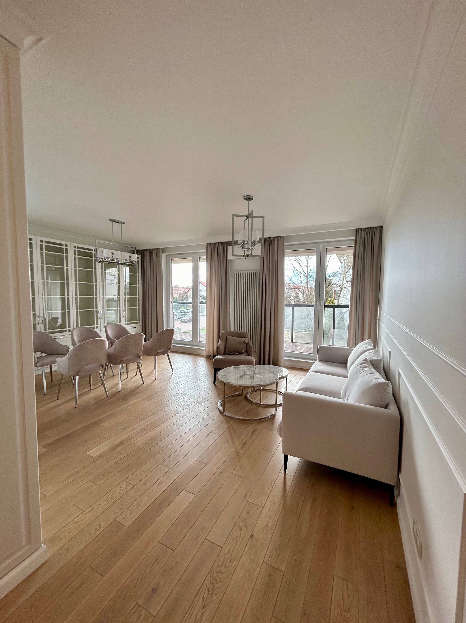 Jasne, przestronne wnętrze po liftingu: parkiet, kremowa sofa, krzesła tapicerowane i dekoracyjne listwy ścienne w eleganckim salonie z dużymi oknami.