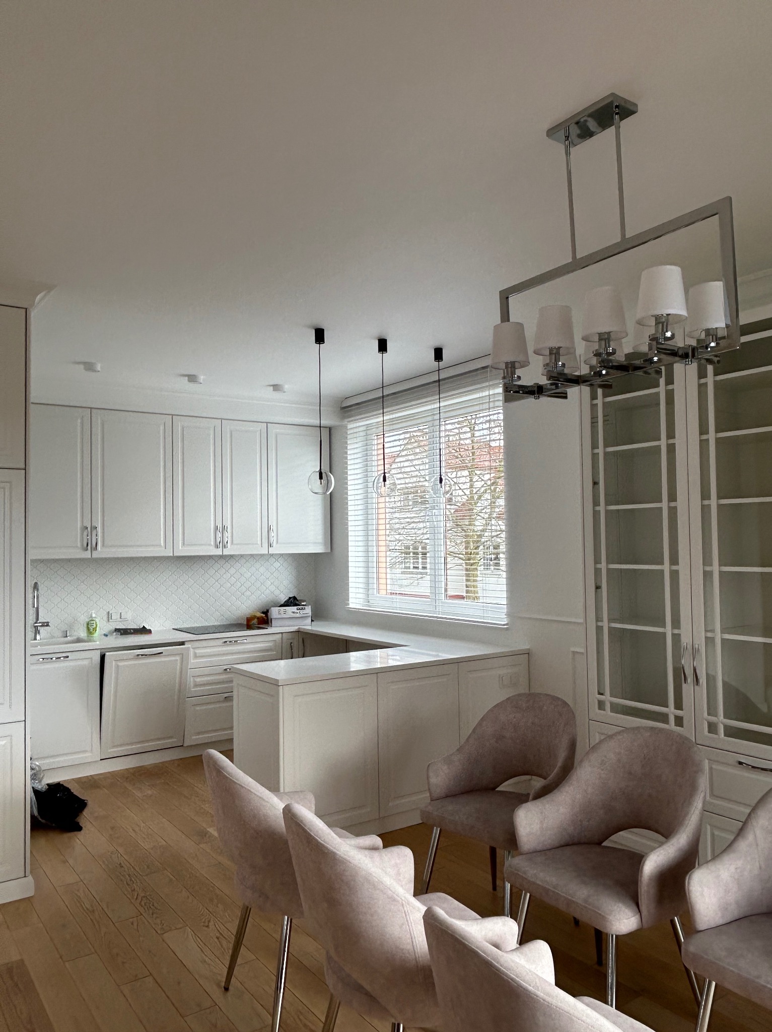 Jasna kuchnia po remoncie z białymi szafkami, wyspą, drewnianą podłogą i krzesłami w kolorze pudrowego różu, oświetlona nowoczesną lampą.