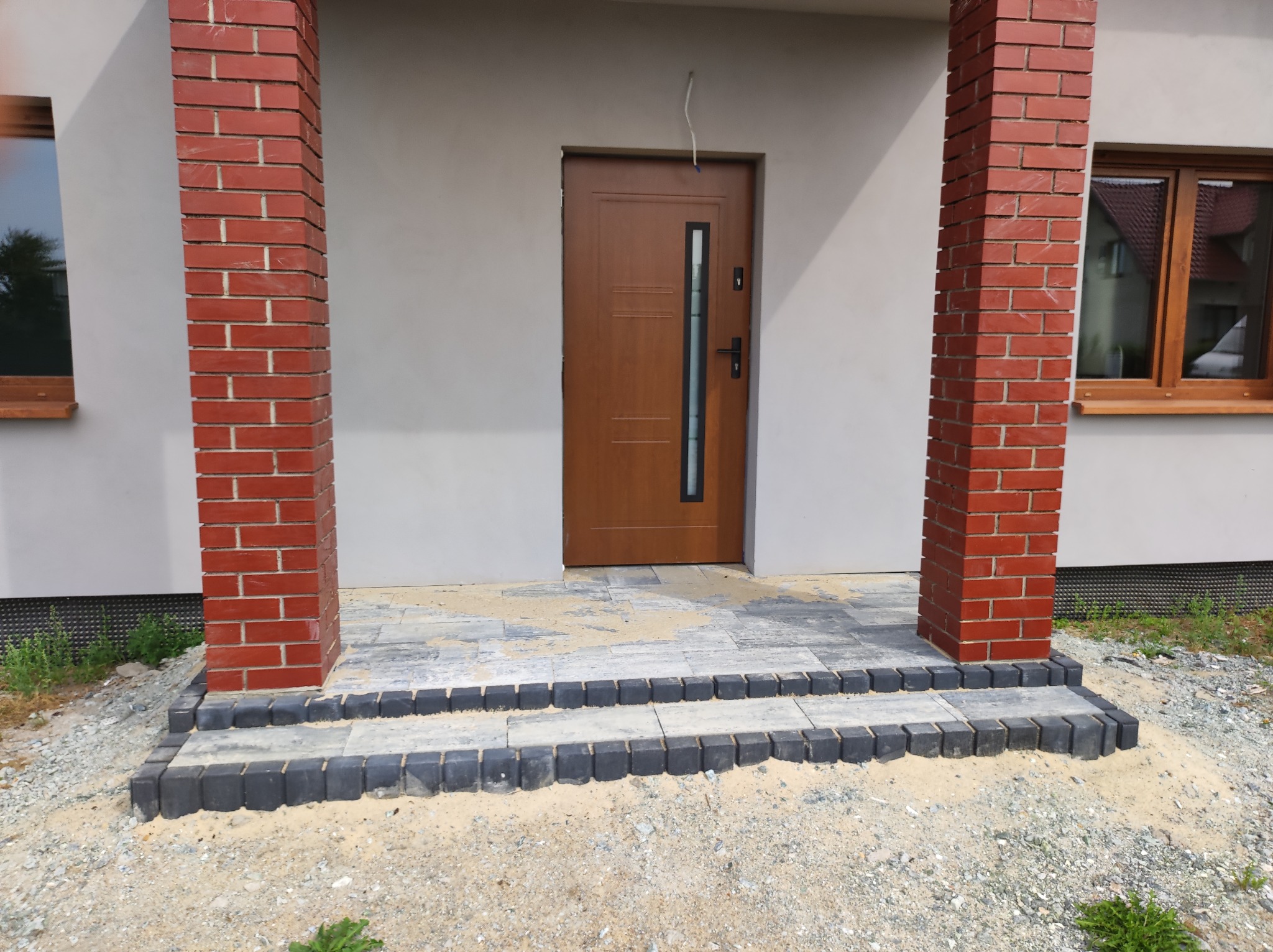 Wejście do domu z nowo ułożoną kostką brukową w odcieniach szarości, obramowane czarnymi obrzeżami i ceglanymi kolumnami, widoczne ślady piasku po montażu.