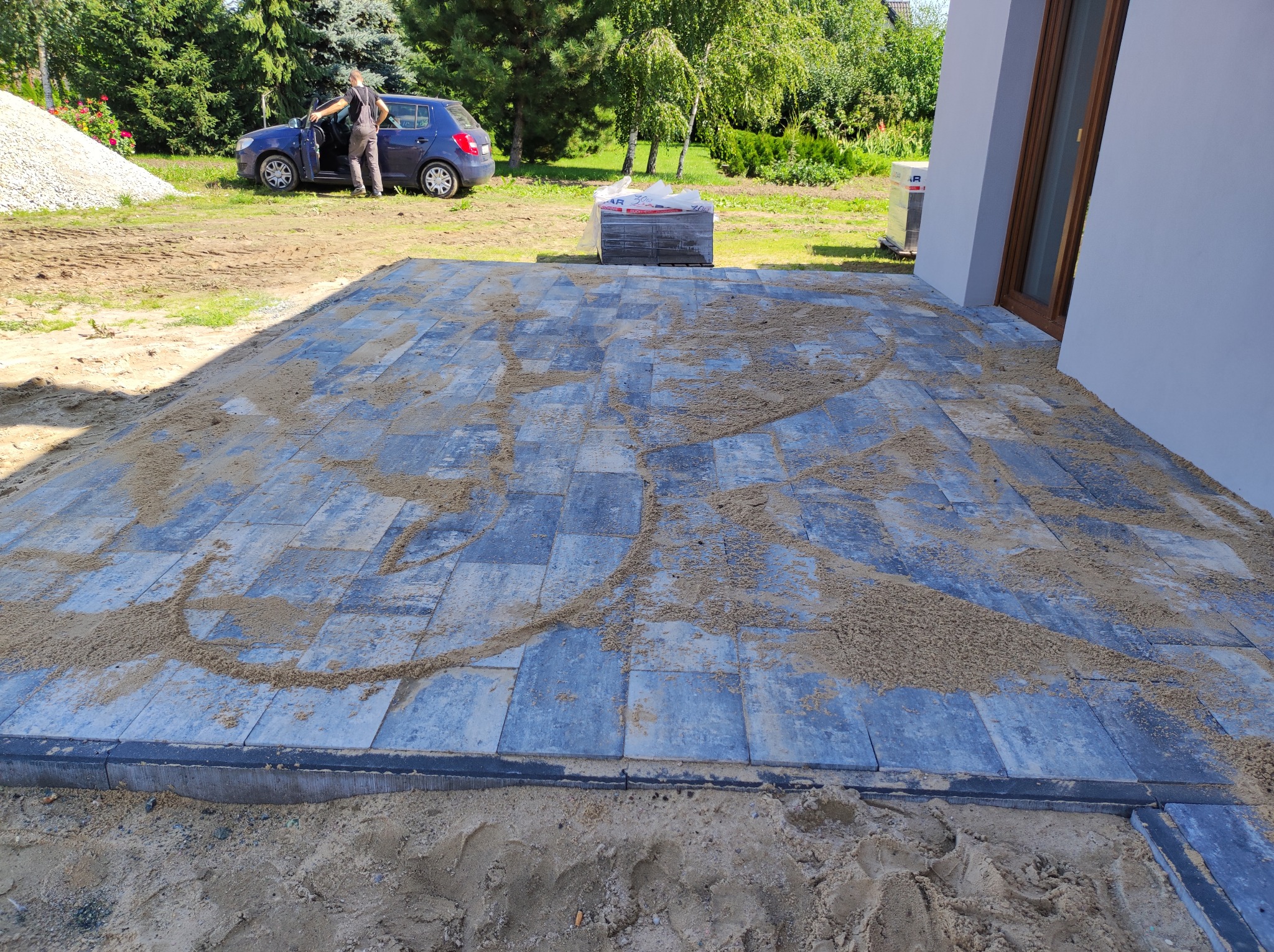 Niedawno ułożona, częściowo zasypana piaskiem, szara kostka brukowa na przygotowanym podłożu, widok z góry, w tle zaparkowany samochód i pracownik w kombinezonie.