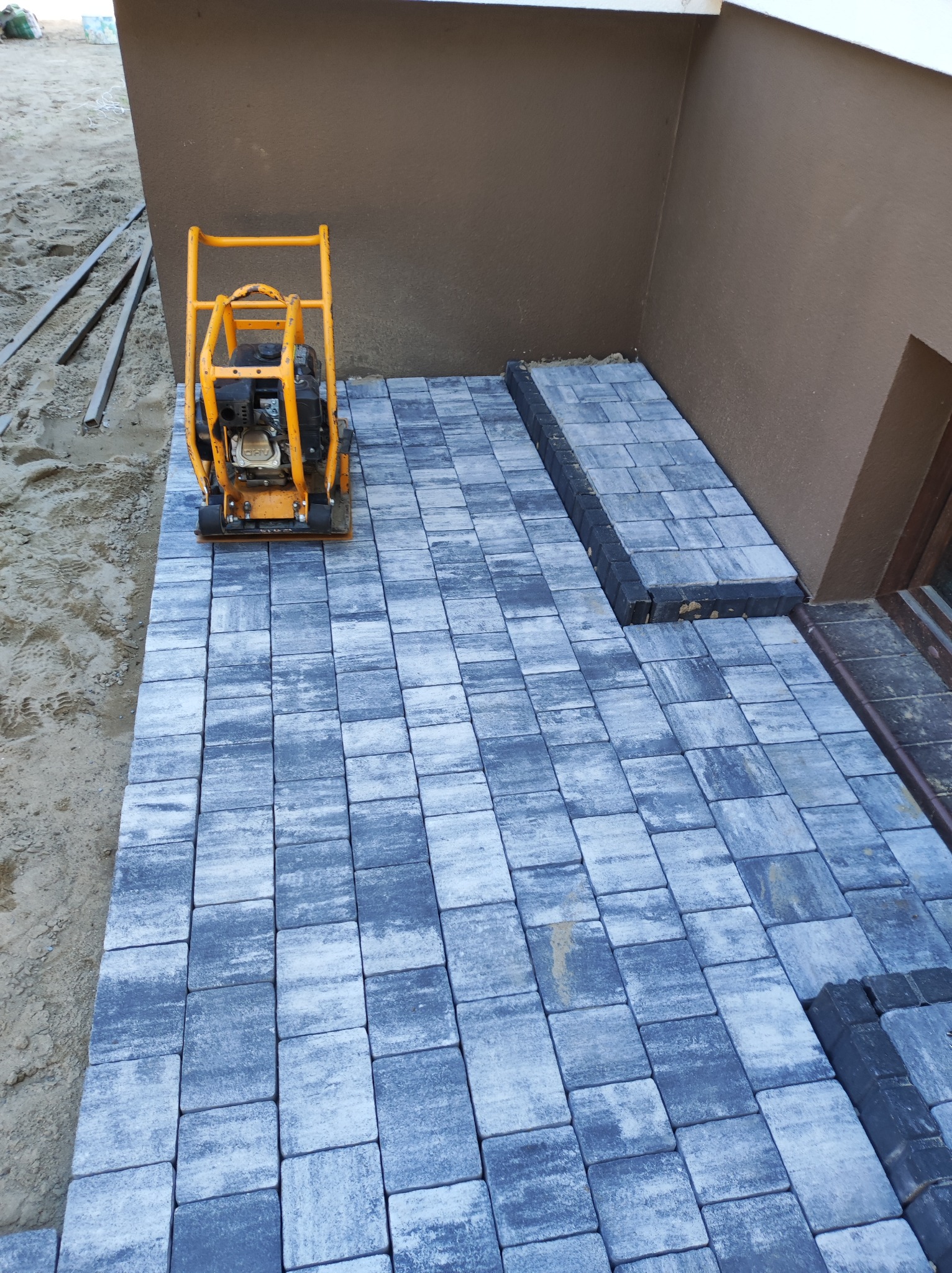 Żółta zagęszczarka obok ułożonej szarej kostki brukowej przy ścianie budynku, z widocznym fragmentem niezabudowanego terenu.