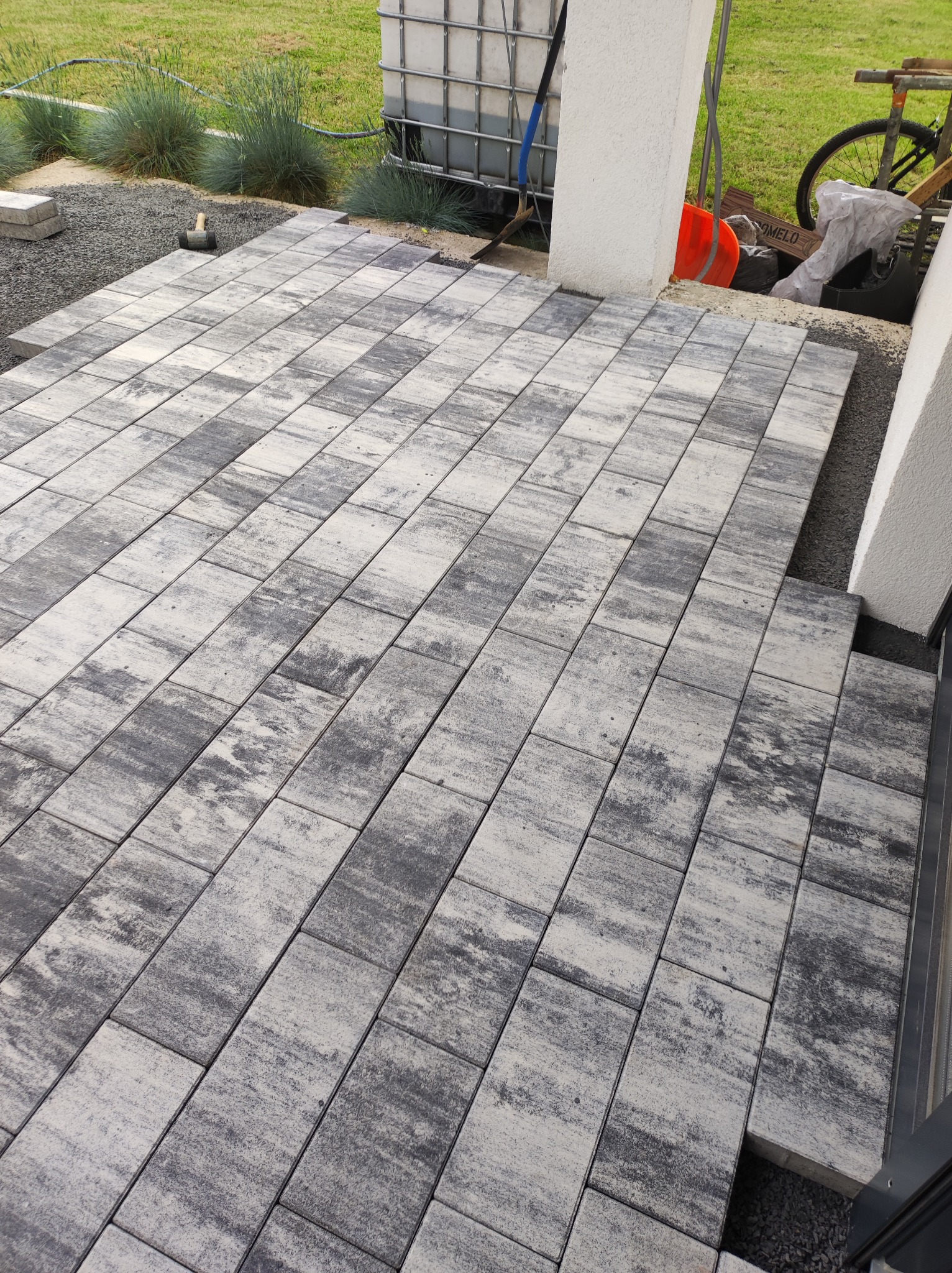 Ułożona nawierzchnia z prostokątnej kostki brukowej w odcieniach szarości i bieli, z widocznym przygotowanym podłożem z grysu, zbiornik na wodę w tle i fragment roweru.