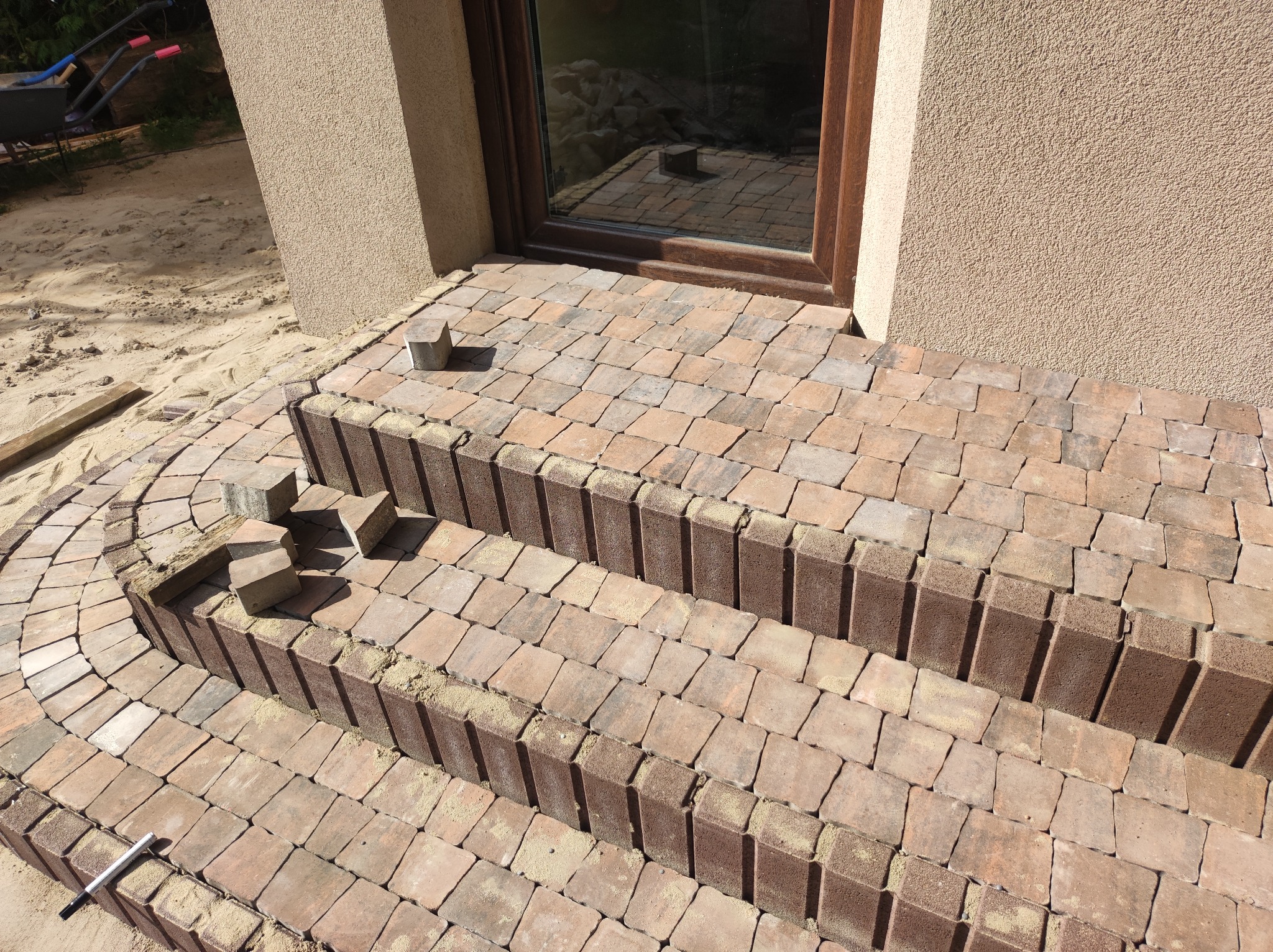 Schody wejściowe do domu w trakcie budowy z kostki brukowej w odcieniach brązu i beżu, widoczne niedokończone fragmenty i narzędzie do układania.