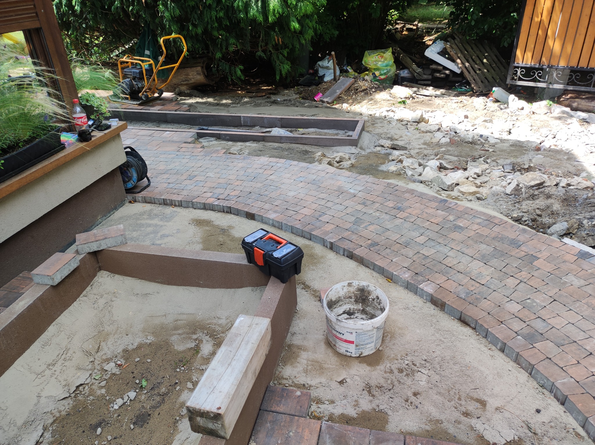 Układanie kostki brukowej: Widok na ścieżkę z kostki brukowej w trakcie budowy, z widoczną warstwą piasku, narzędziami (skrzynka narzędziowa, wiadro) i przygotowanym miejscem na rabatę z kostki.